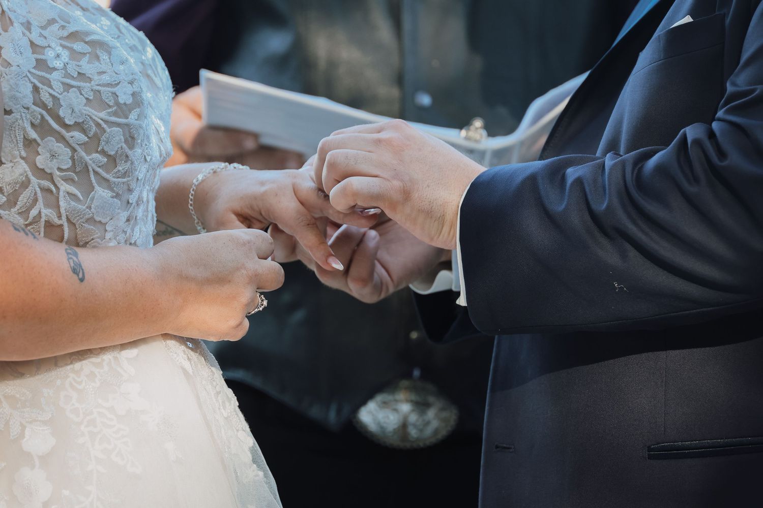 Close up of hands exchanging wedding rings during marriage ceremony with white lace dress and dark suit visible.