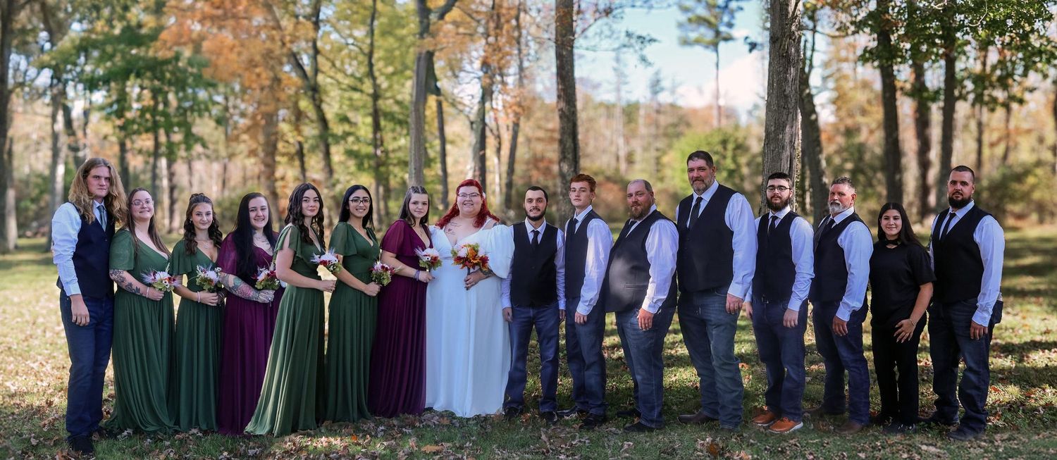 Large wedding party poses outdoors in burgundy and navy attire during autumn with colorful fall foliage in background.