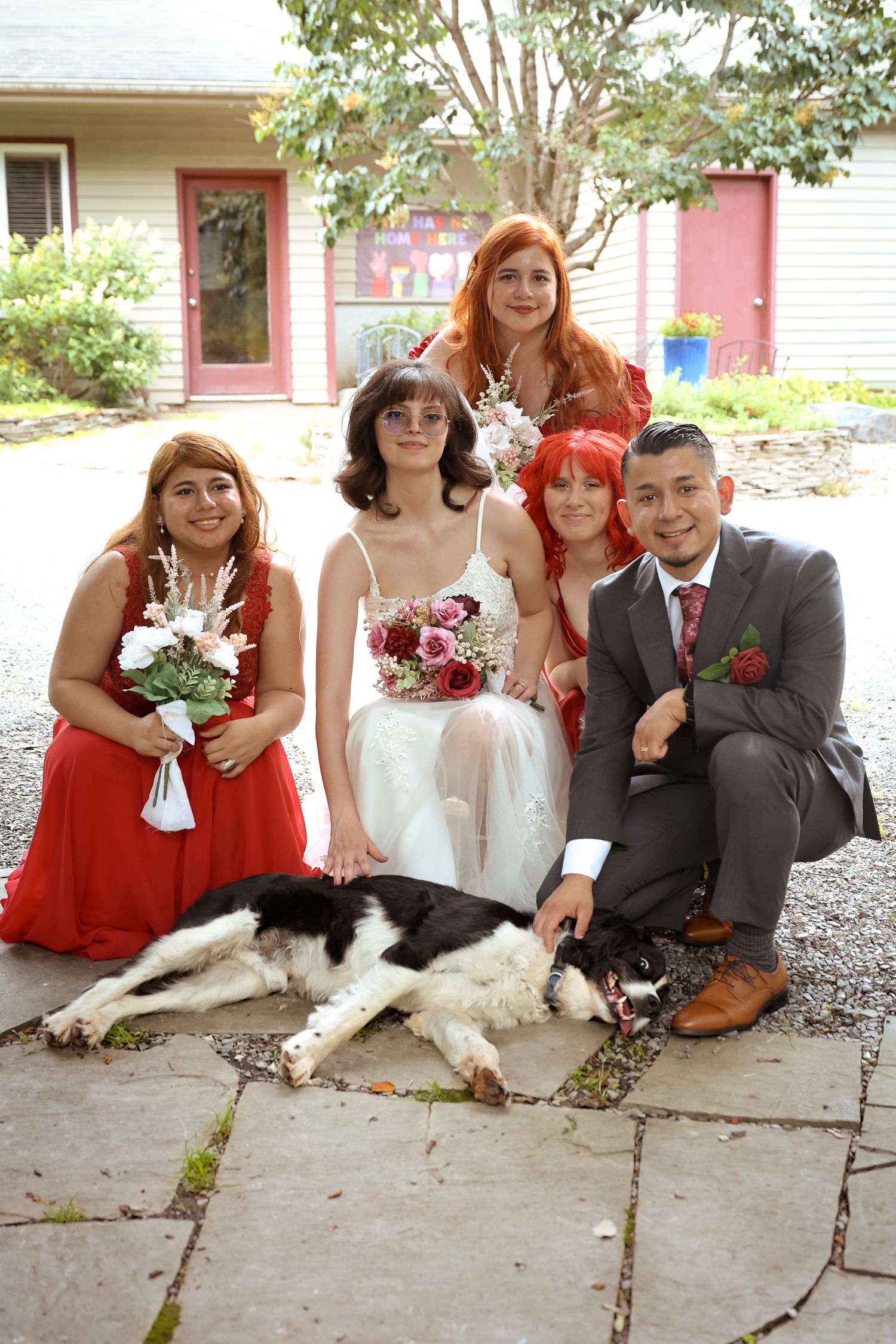 Wedding party in red dresses and gray suit pose outdoors with a large black and white dog laying on stone patio.