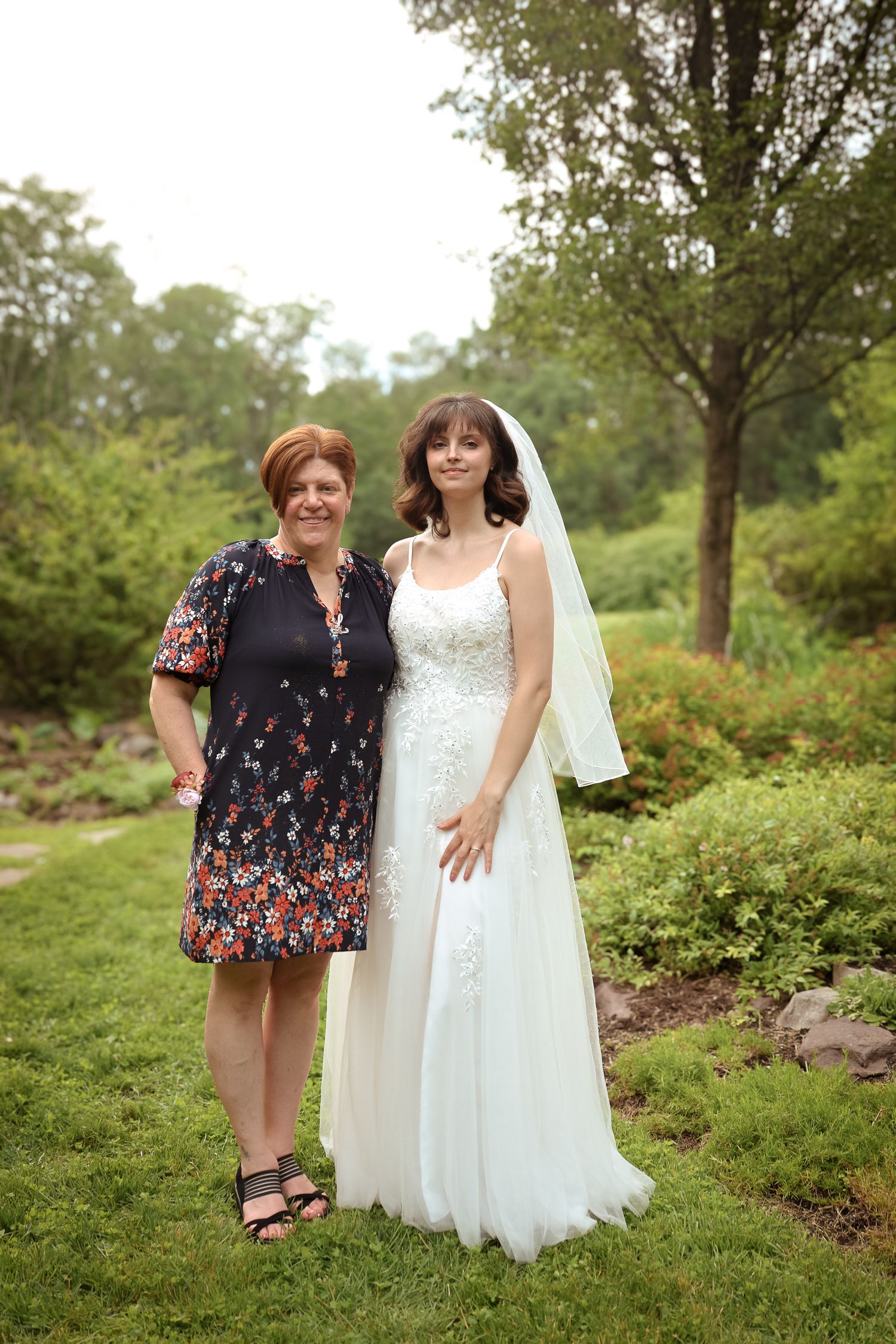 Two people pose together outdoors in a garden setting with lush greenery and trees in the background.