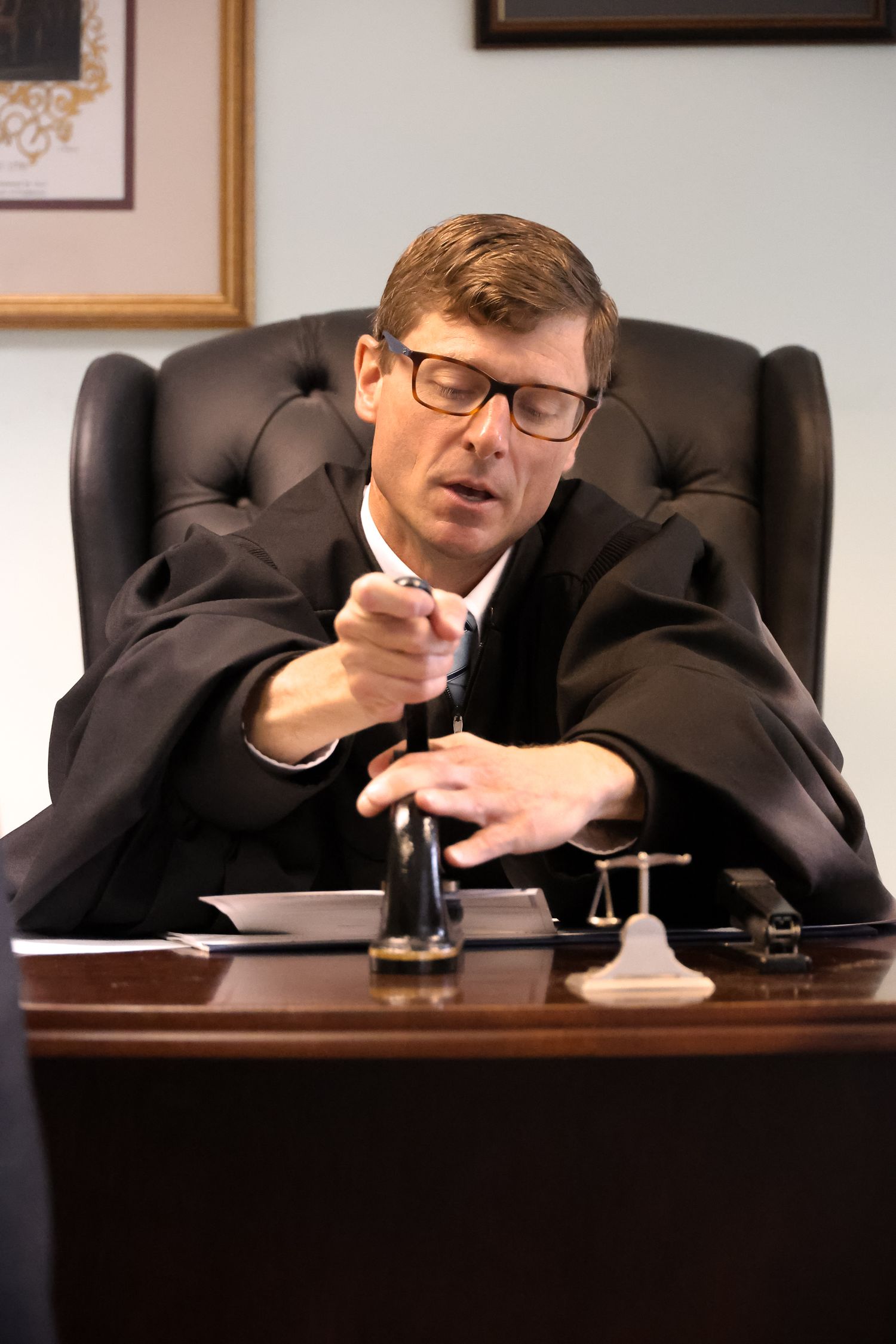 Judge in black robes and glasses yawns while sitting at bench in courtroom.