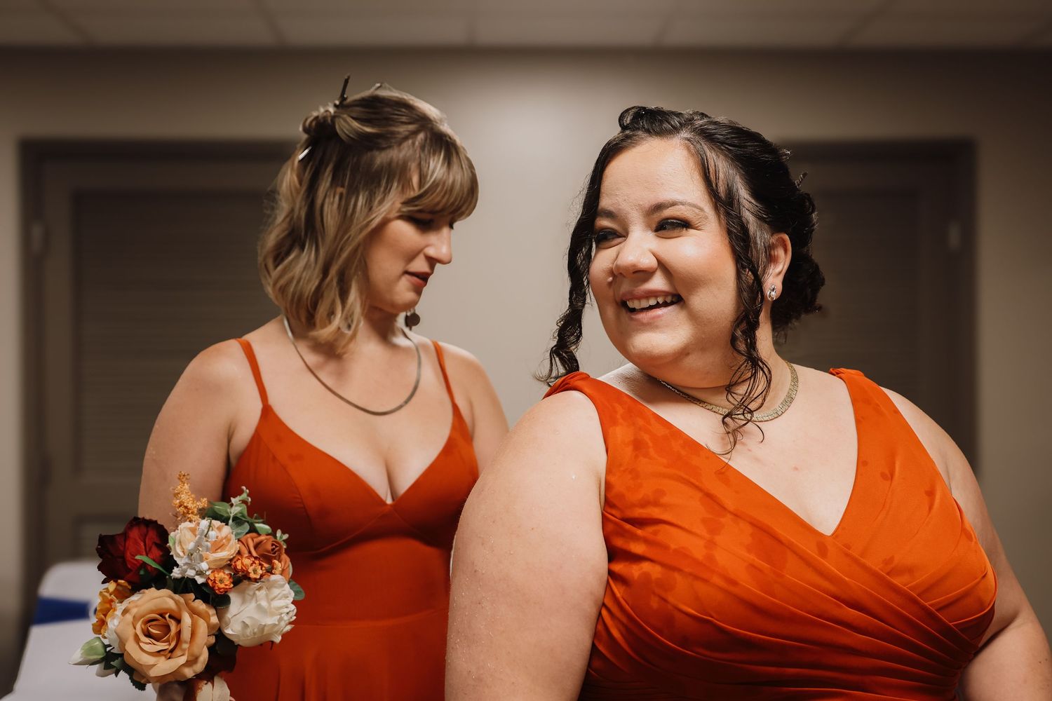 Bridesmaids in vibrant orange dresses share a candid laughing moment during a wedding celebration.