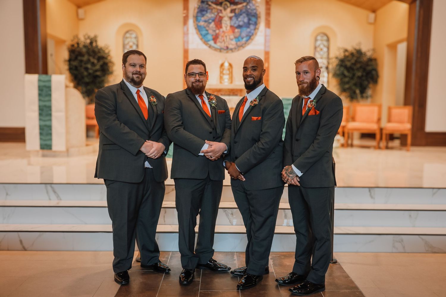 A wedding party in dark suits stands in a warm-lit church sanctuary with wooden pews and decorative greenery.