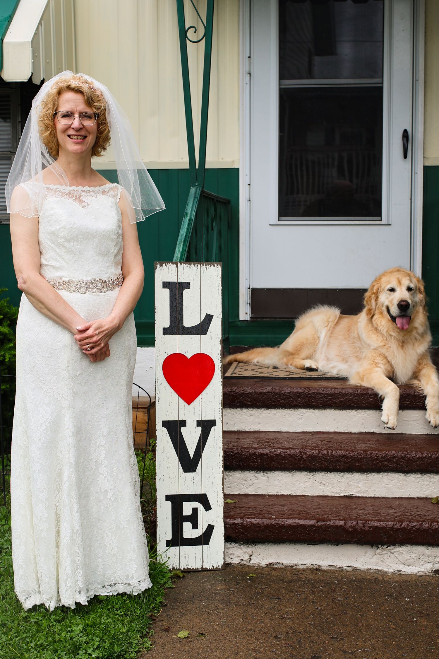 A bride in a white lace wedding dress stands next to a LOVE sign on porch steps while a golden retriever rests nearby.