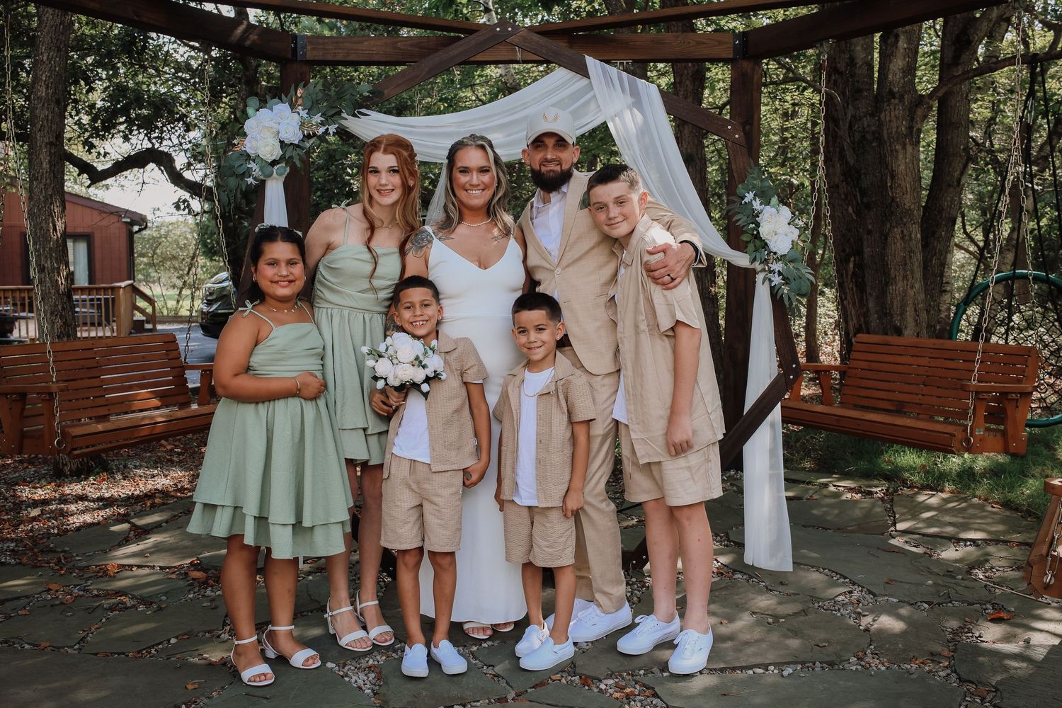 A group in coordinated pastel outfits poses together outdoors near trees and wooden structures.