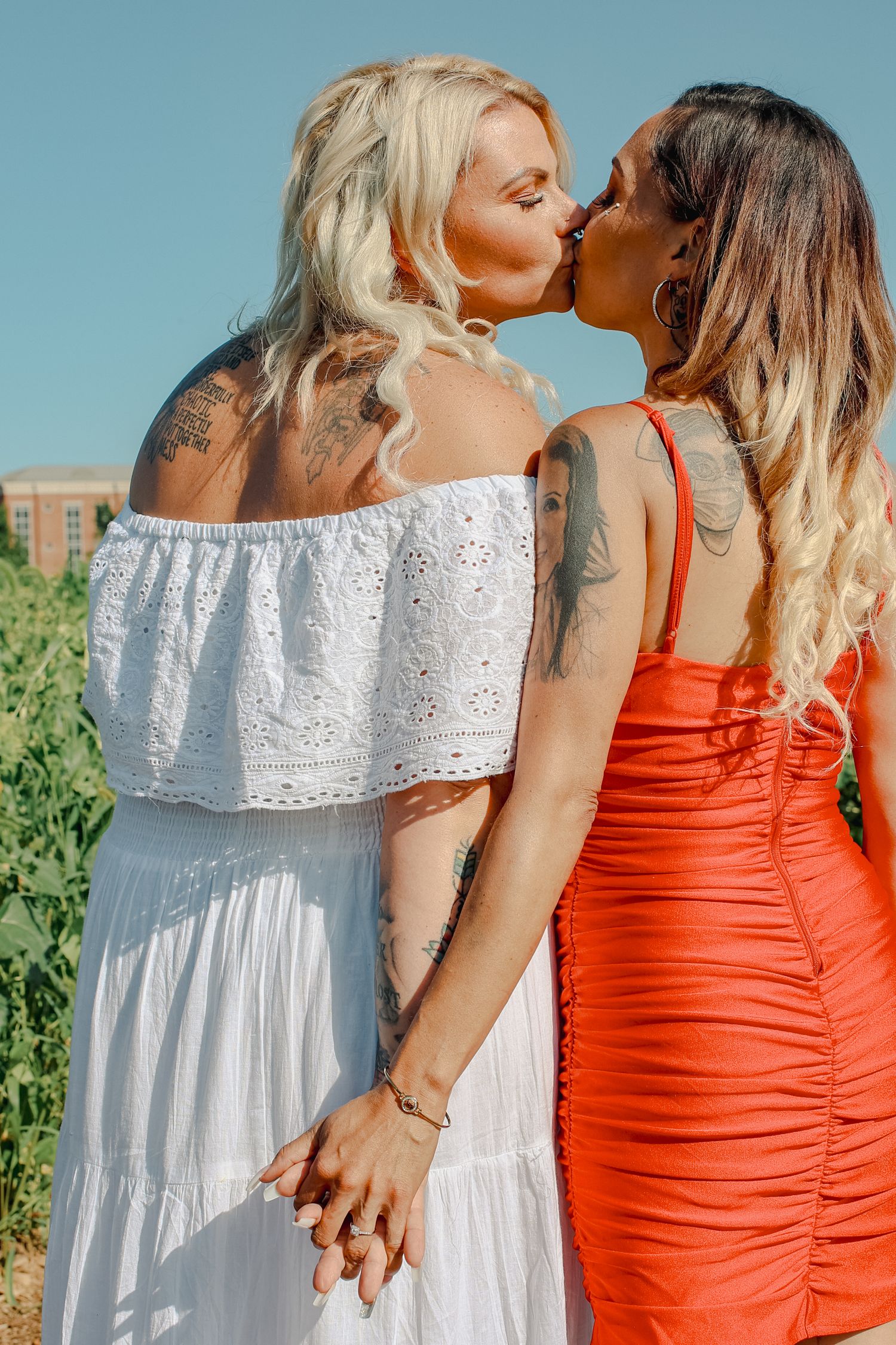 Two people in contrasting white and orange summer dresses embrace outdoors against a blue sky background.