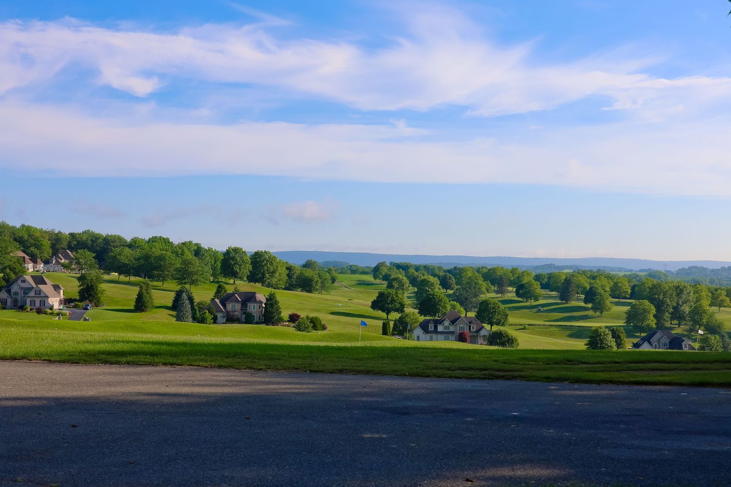 Rolling green hills stretch across a scenic rural landscape under a bright blue sky with scattered white clouds.