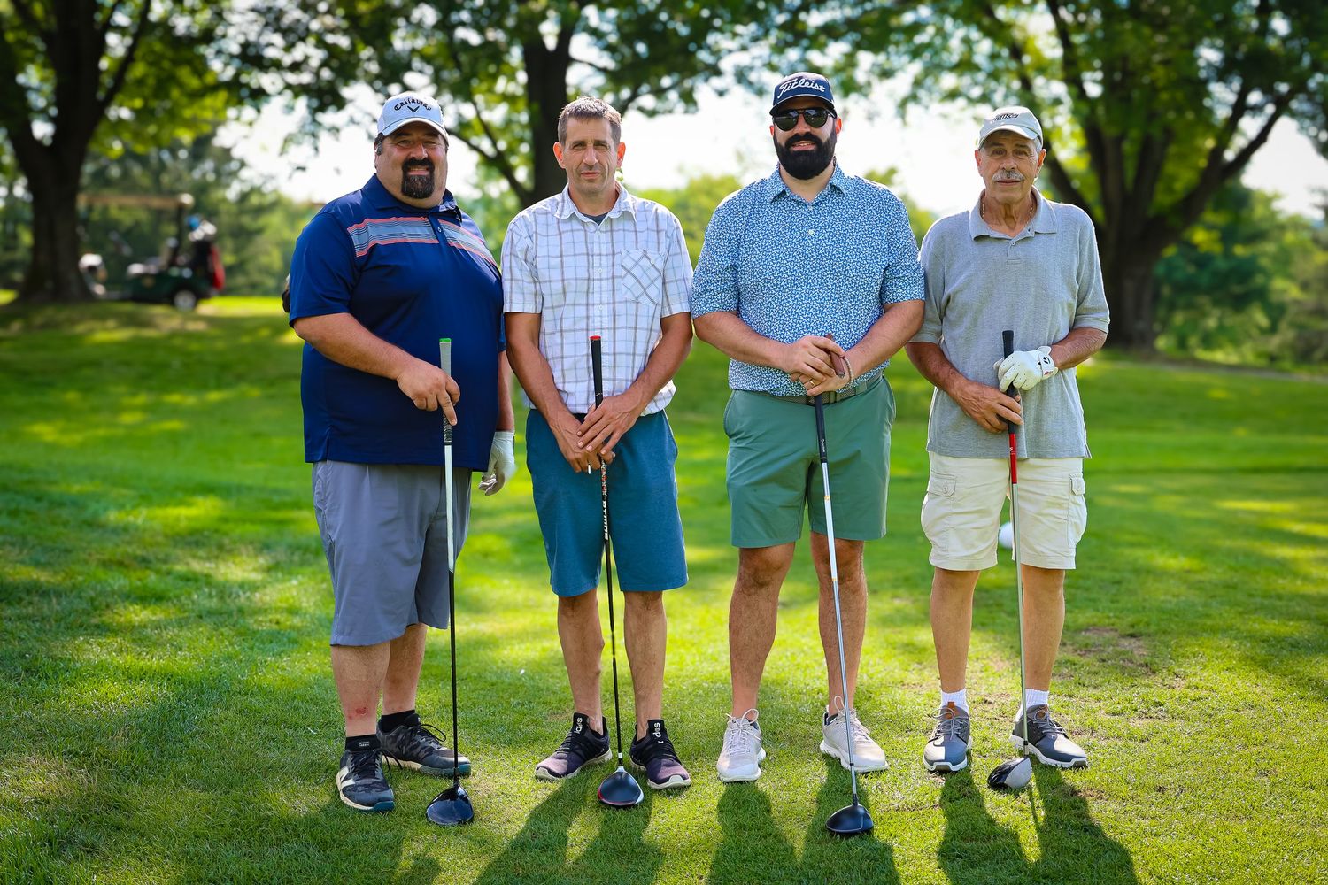 Four people stand together on a sunny golf course holding golf clubs and wearing casual attire.