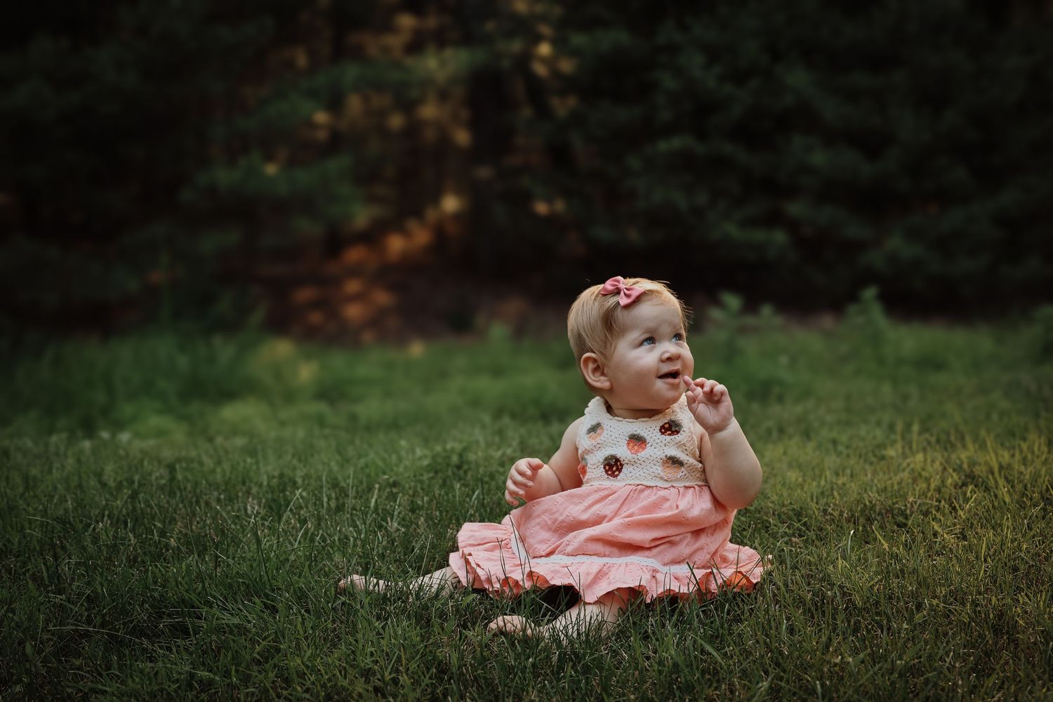 A small child in a pink dress sits on green grass during an outdoor photo session.
