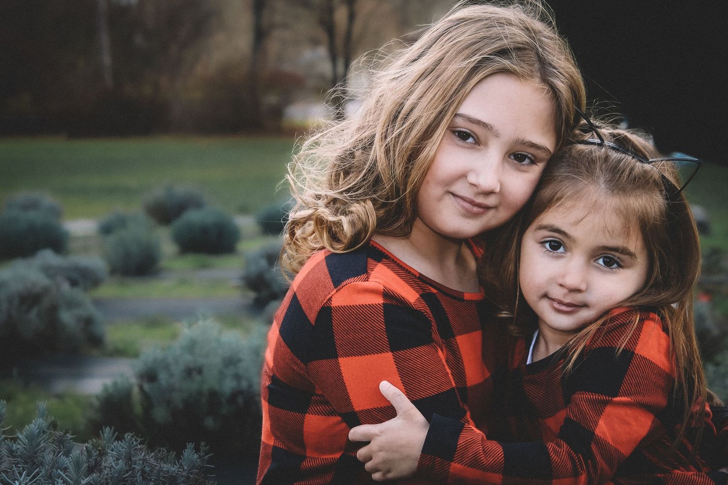 Two people in matching red and black buffalo plaid jackets embrace while sitting outdoors in a garden setting.