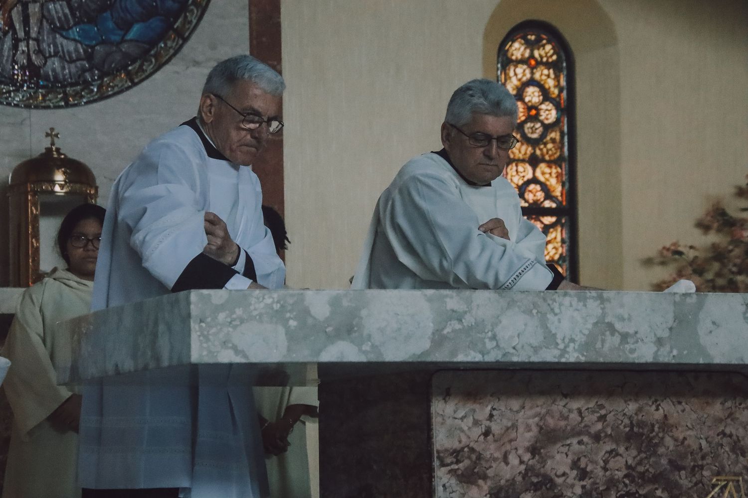 Two clergy members in light blue robes stand at a marble altar during a religious service.