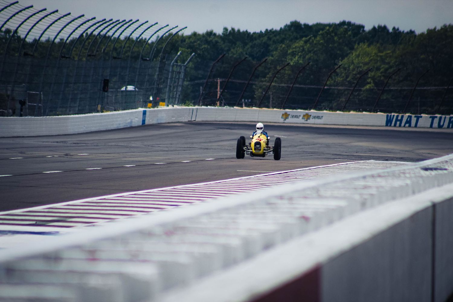 Vintage Formula 1 race cars line up on a gray racetrack with trees and cloudy skies in the background.