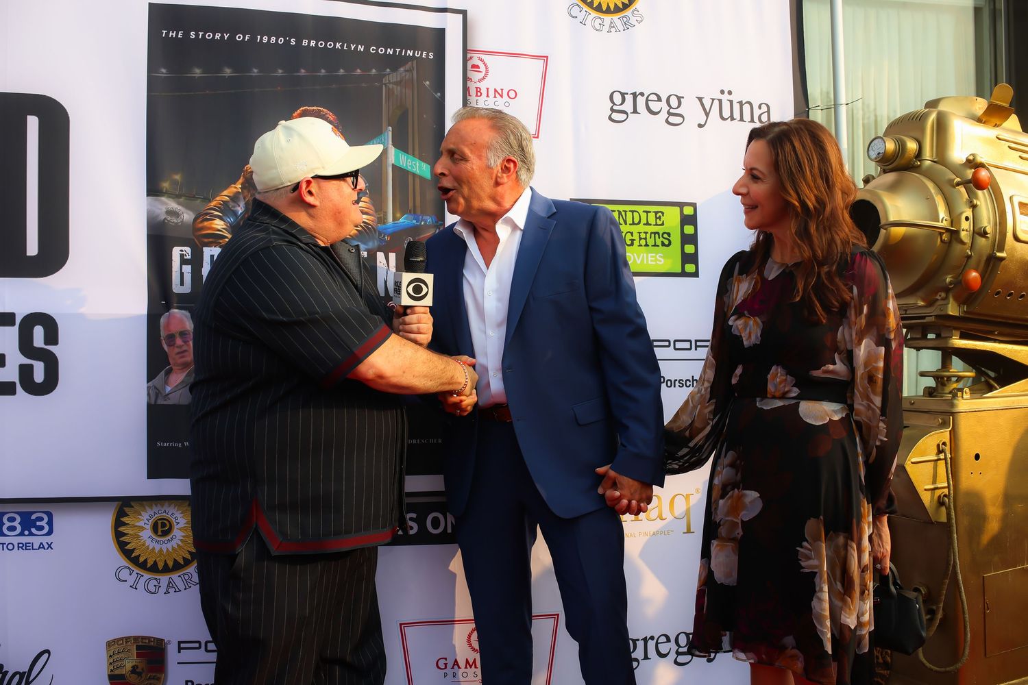 Two people shake hands at a red carpet event with promotional banners and movie posters in the background.