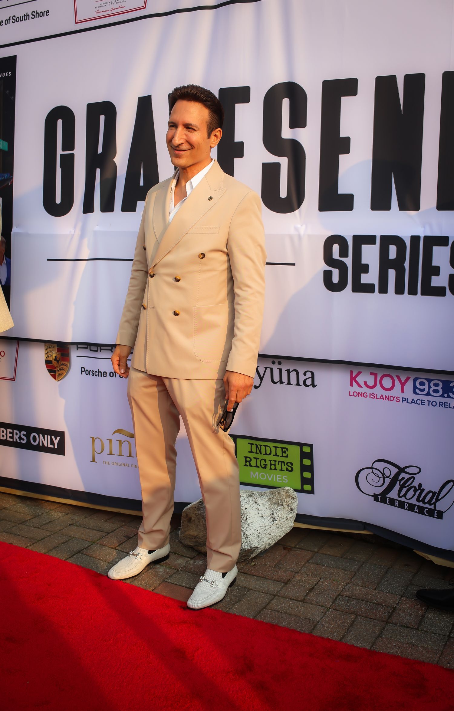 A person in a cream-colored suit and white shoes poses on a red carpet at an event.