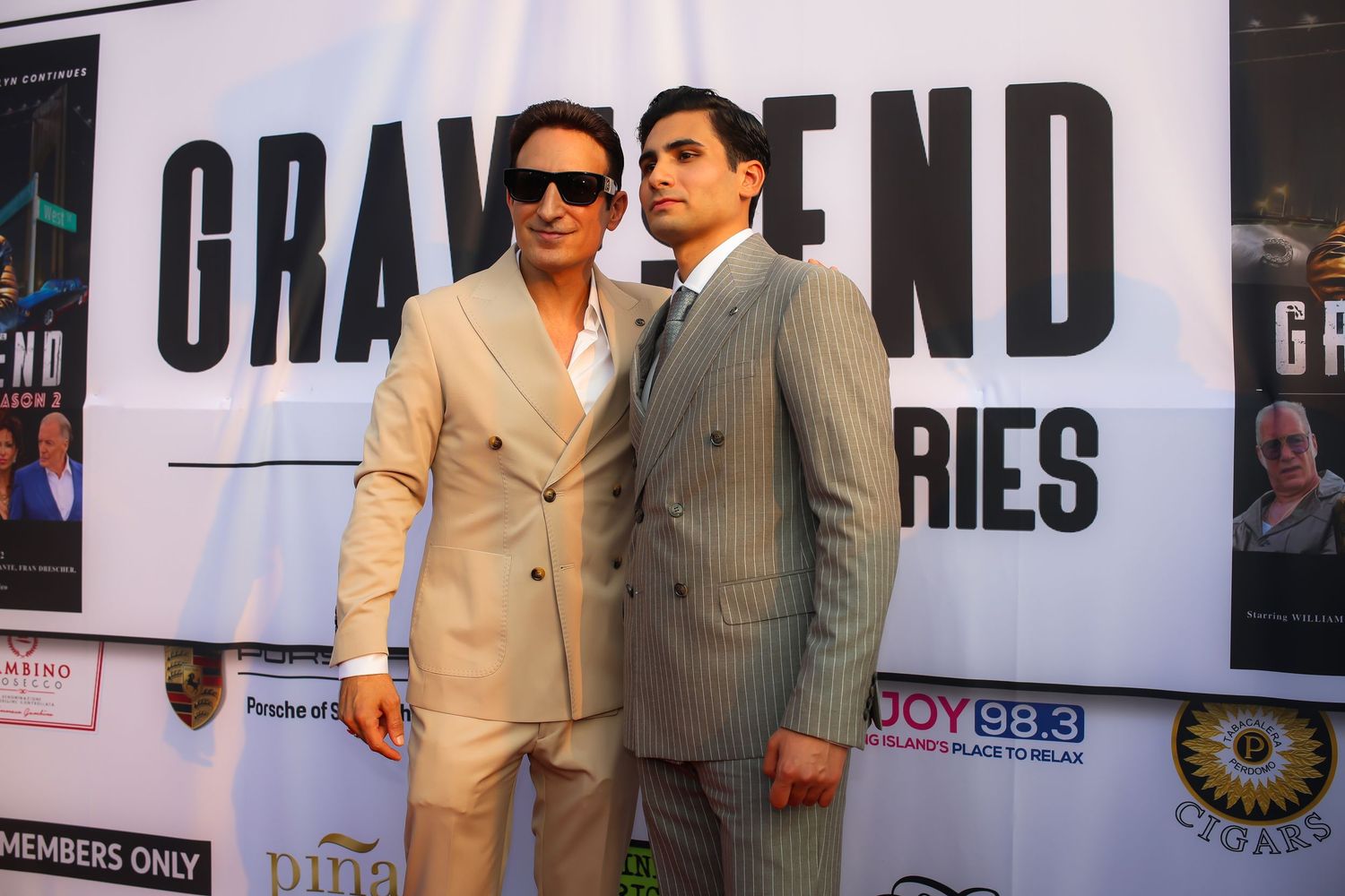 Two people in formal attire pose together at an event with promotional banners visible in the background.
