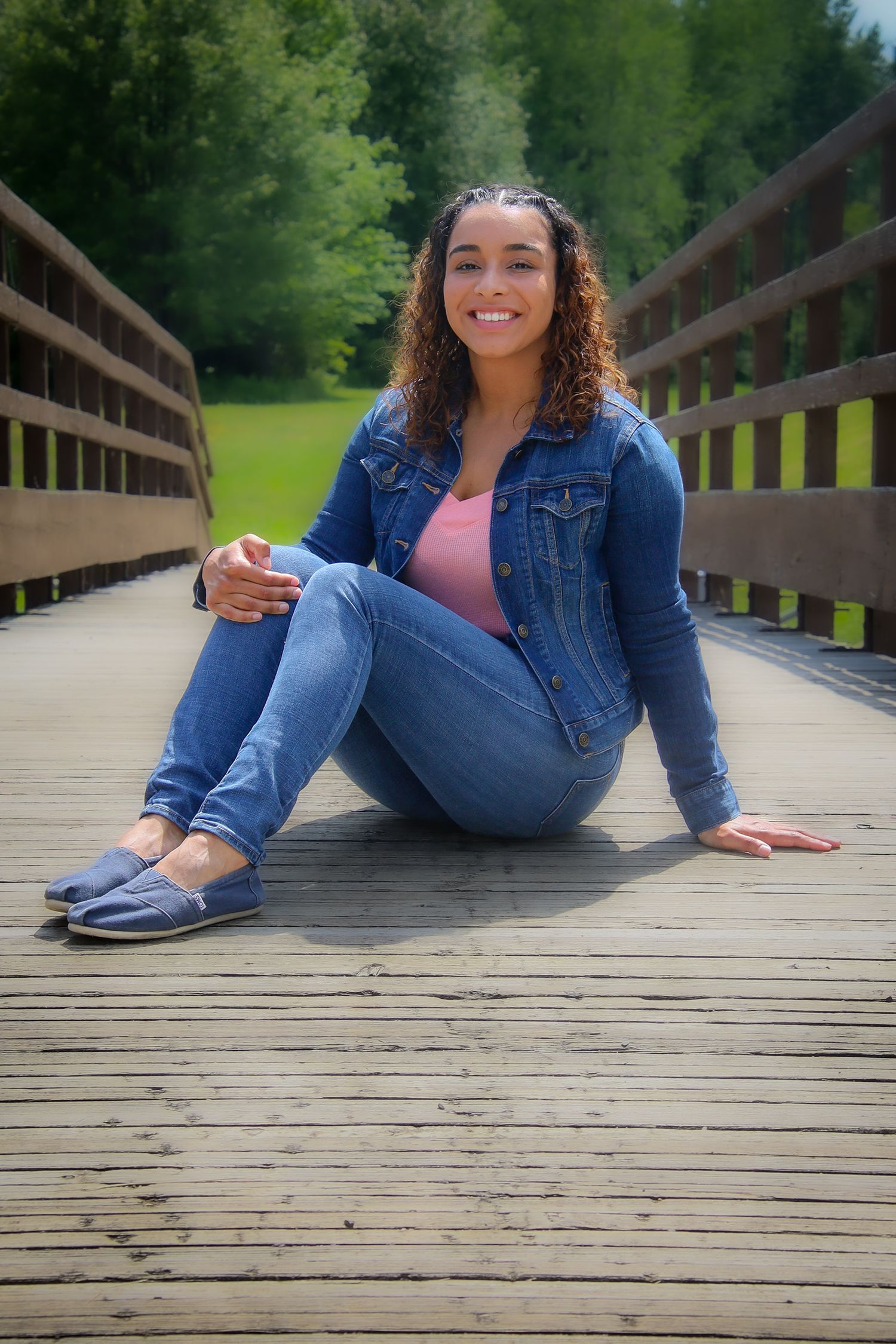 Person in denim jacket and jeans sitting casually on wooden boardwalk with green forest backdrop.