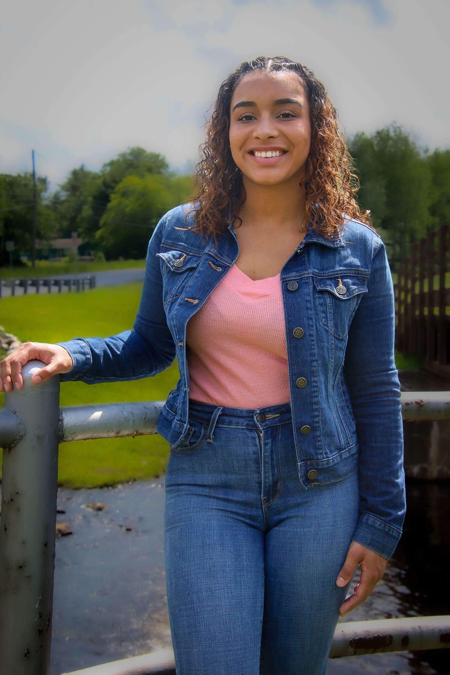 A person in a denim jacket and pink top poses outdoors near a waterfront on a sunny day.