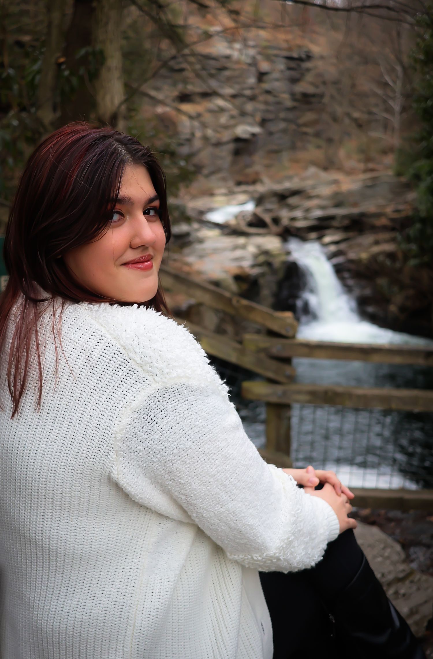 A person in a cozy white sweater poses near a scenic waterfall on a winter day.