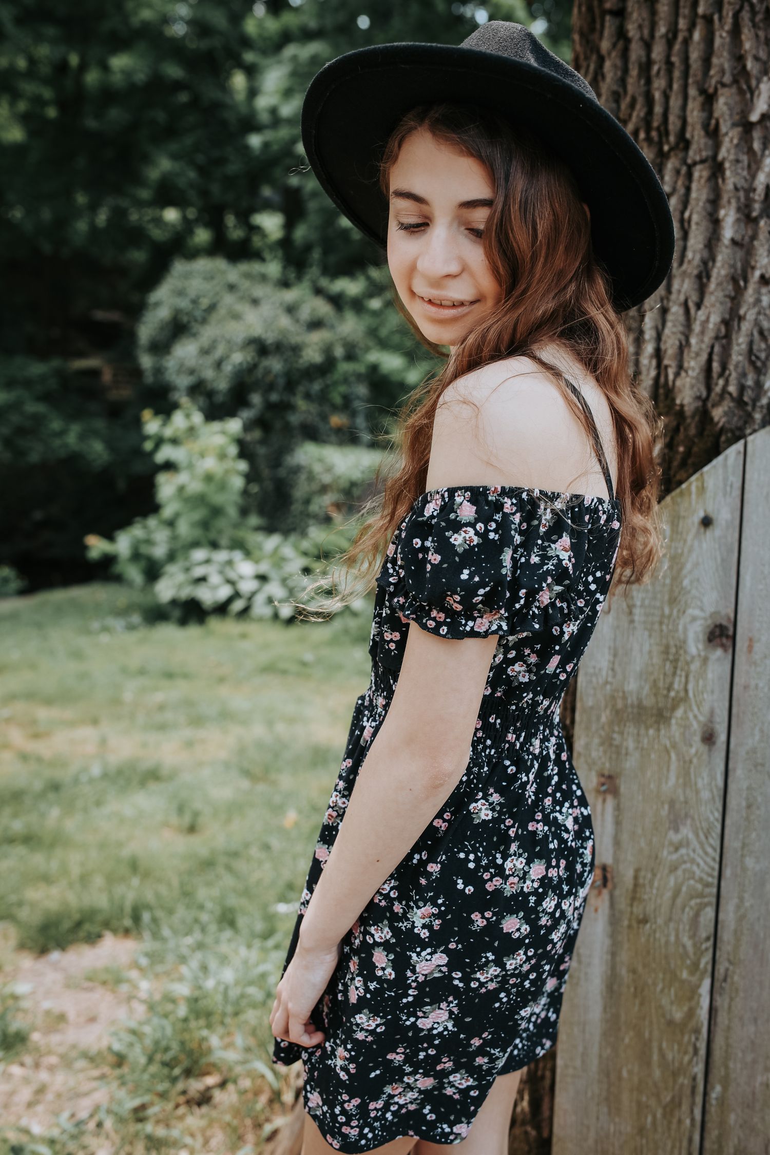 A person in a dark floral off-shoulder dress and wide-brimmed hat poses against a tree in a garden setting.