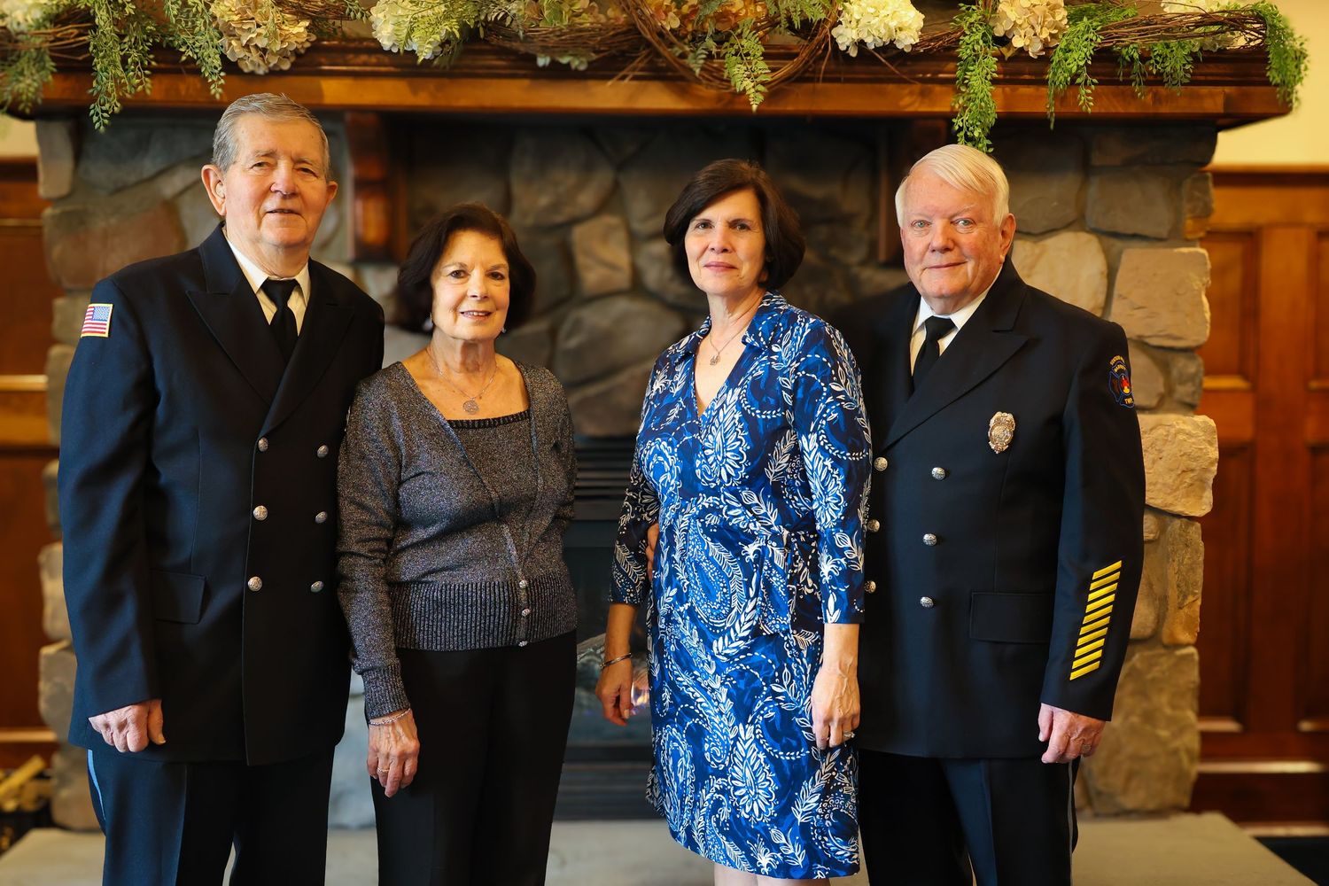 A group of people in formal attire pose together at a social gathering with a stone wall backdrop.