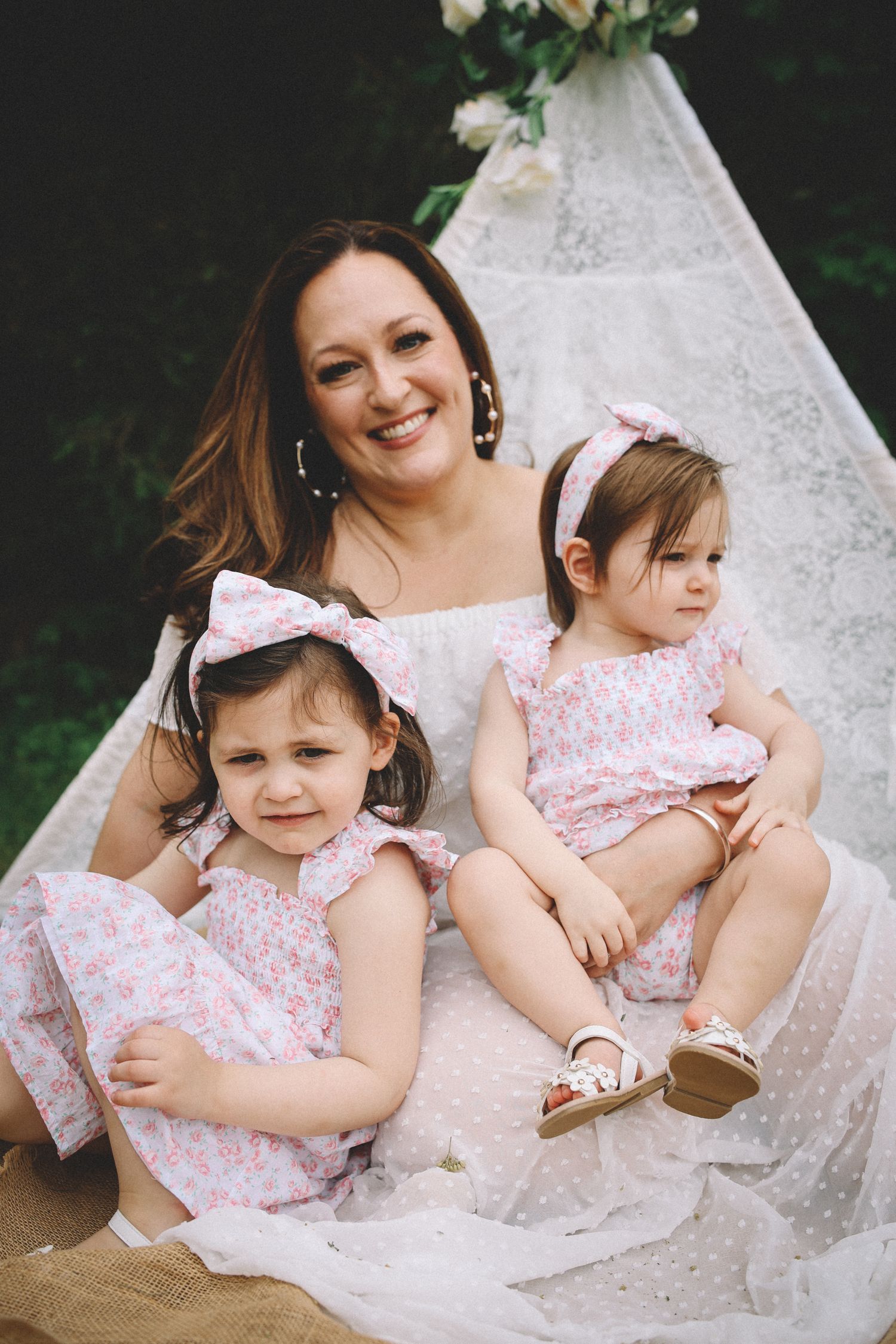 Three people wearing matching pink floral outfits sit together on white fabric outdoors for a family photo.