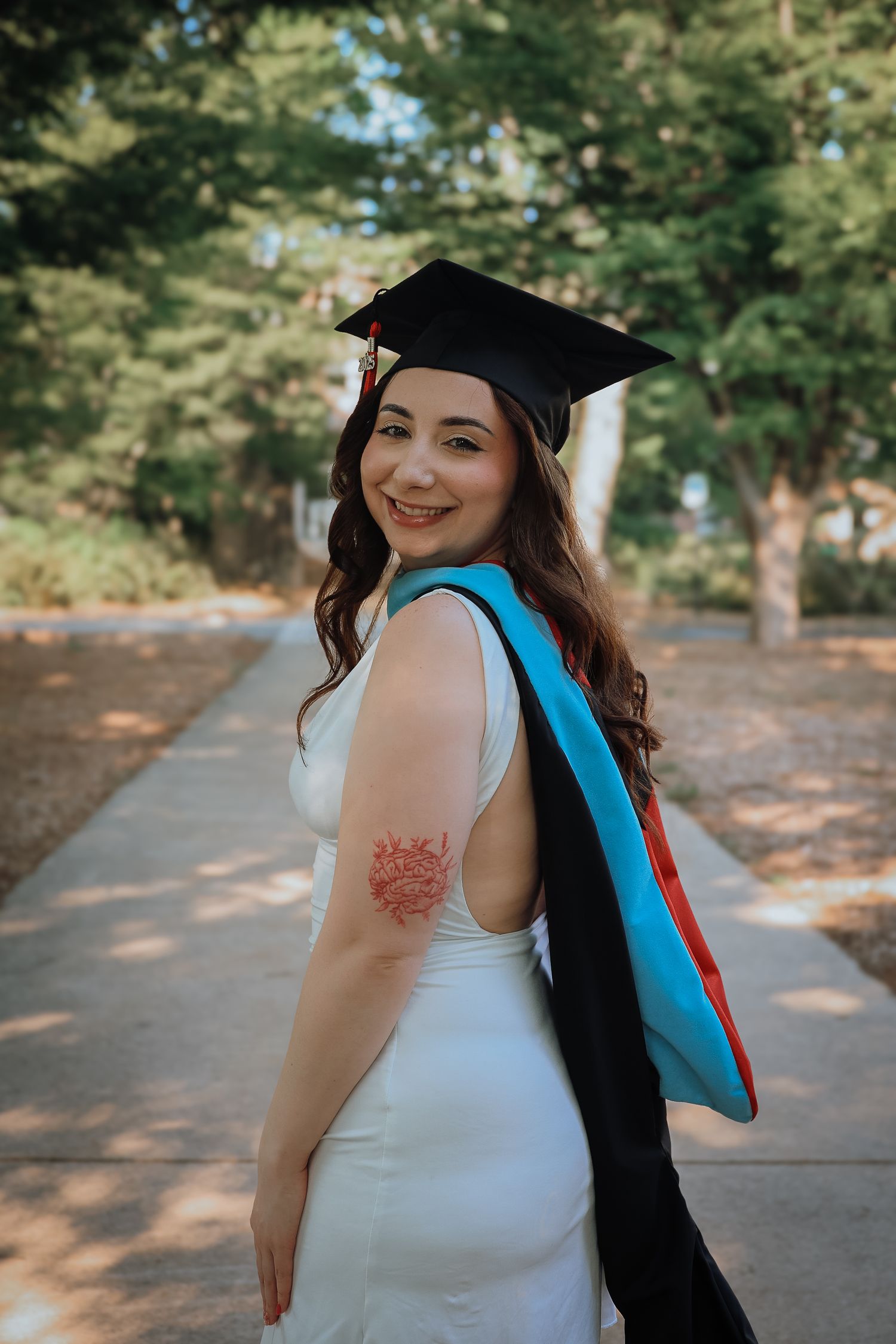 Graduate in white dress and cap smiles while posing on campus pathway.