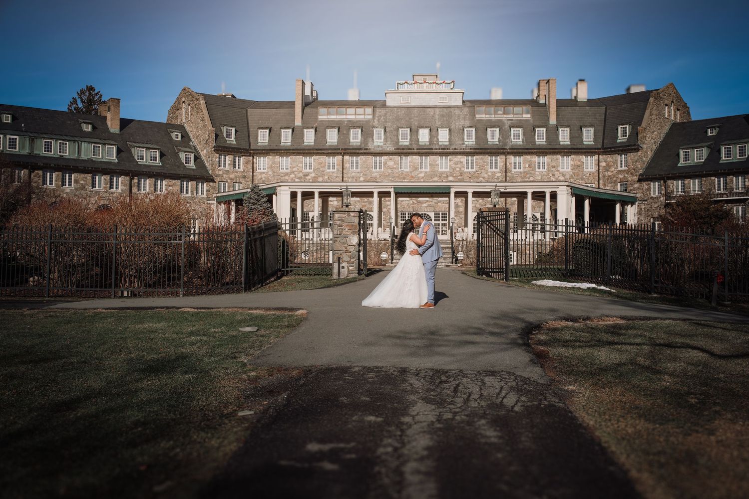 A couple in wedding attire poses in front of a grand historic mansion with symmetrical architecture and manicured grounds.
