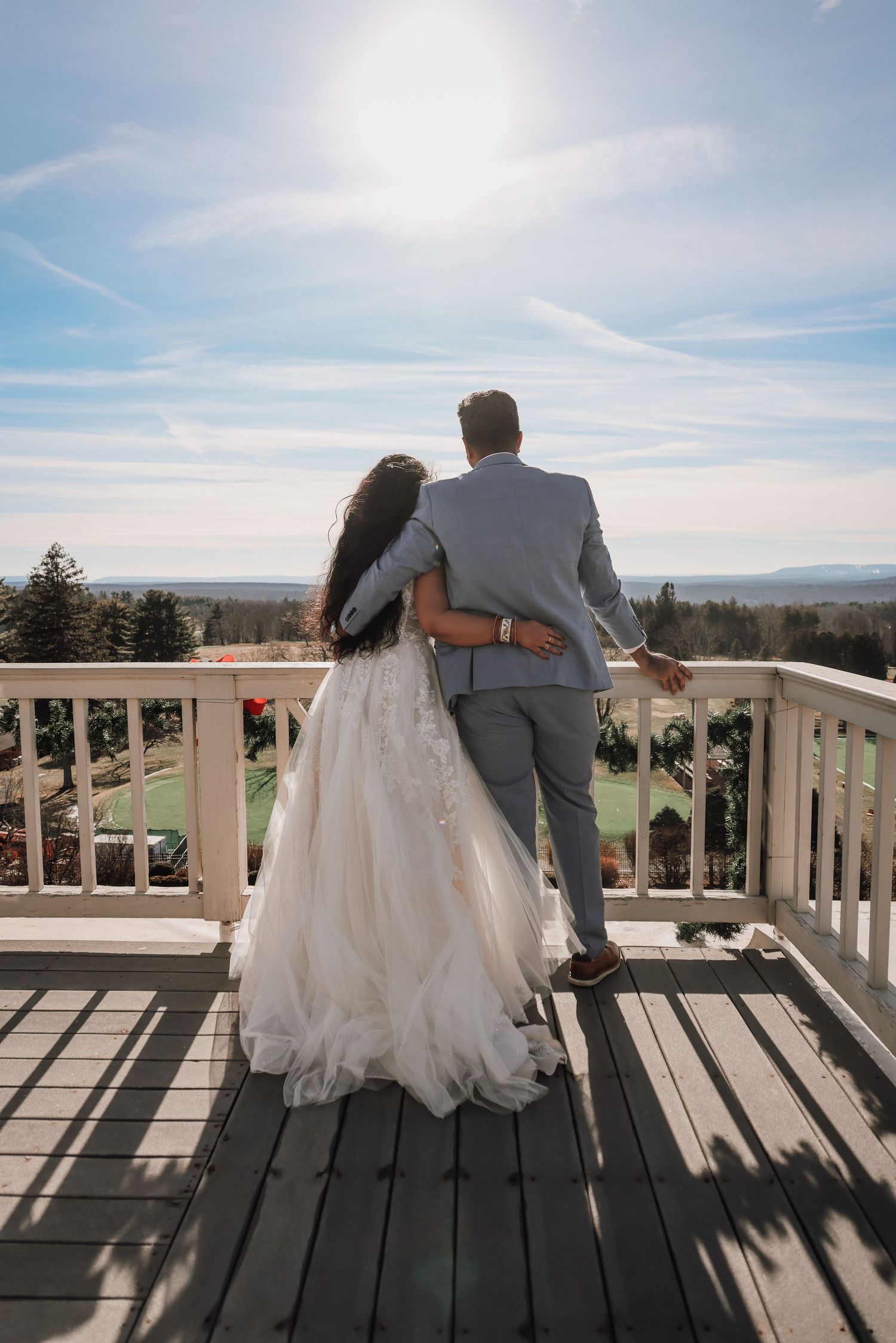 A couple in wedding attire sits together on a wooden deck overlooking a scenic mountain landscape on their special day.