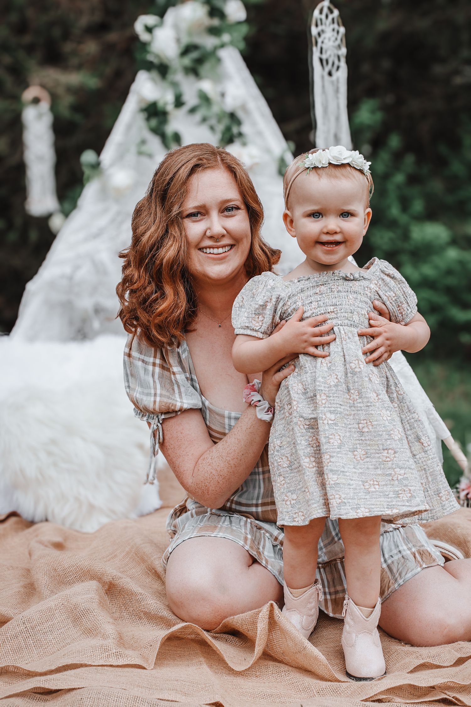 Mother and baby girl in matching plaid outfits pose on blanket with teepee decorations in outdoor setting.