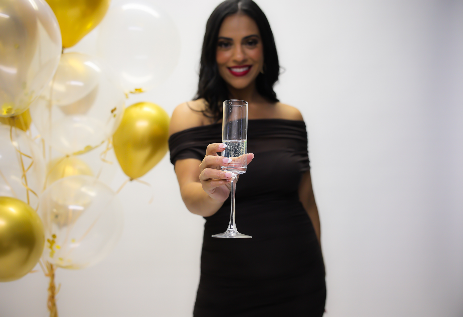 Person in a black dress raises a cocktail glass in celebration with gold balloons in the background.