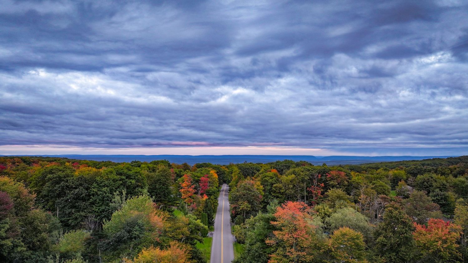 Panoramic view of autumn trees and dramatic cloudy sky over rolling hills with a lighthouse in the distance.