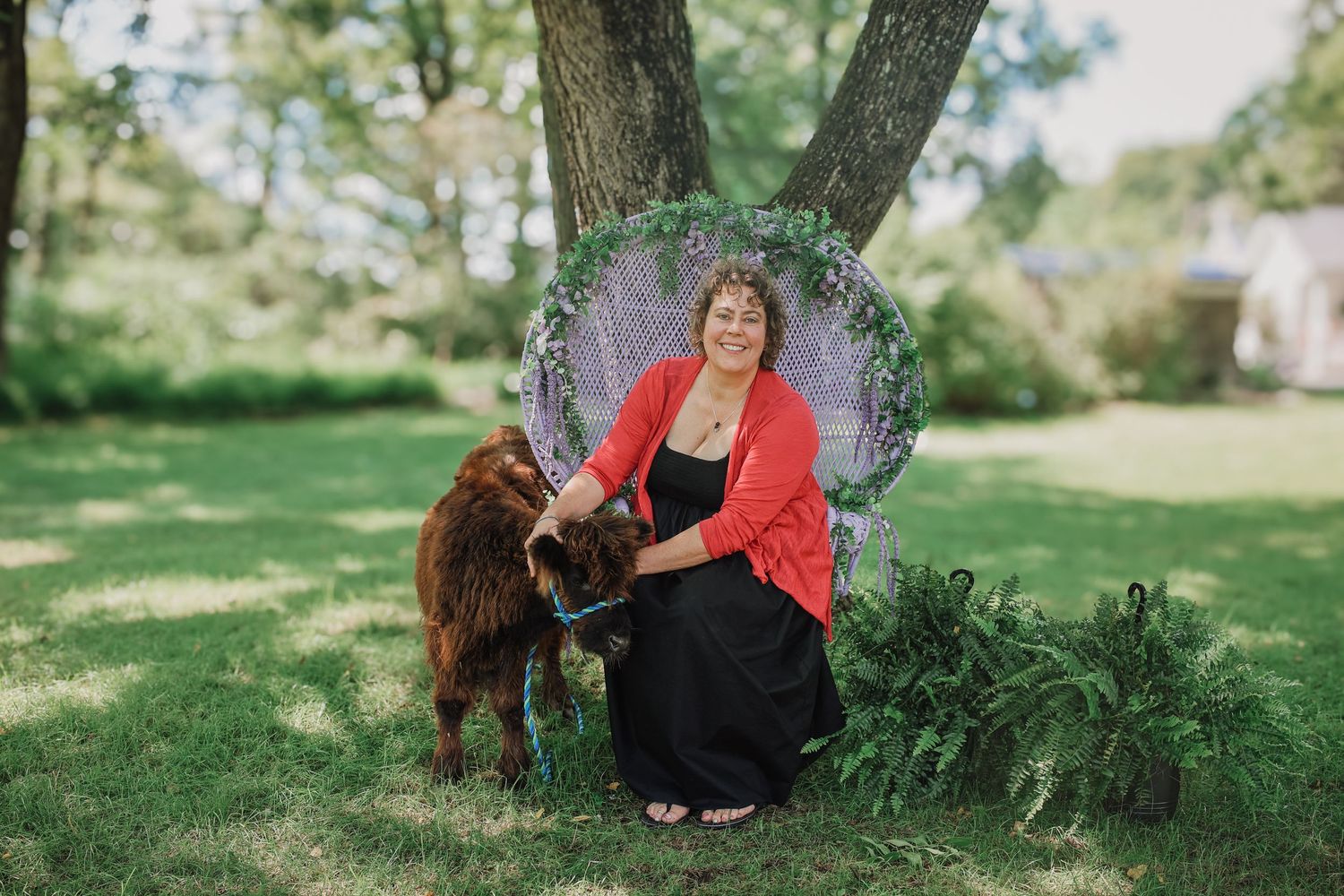 A person in a red dress sits on the grass with a brown dog in a sunny outdoor setting.