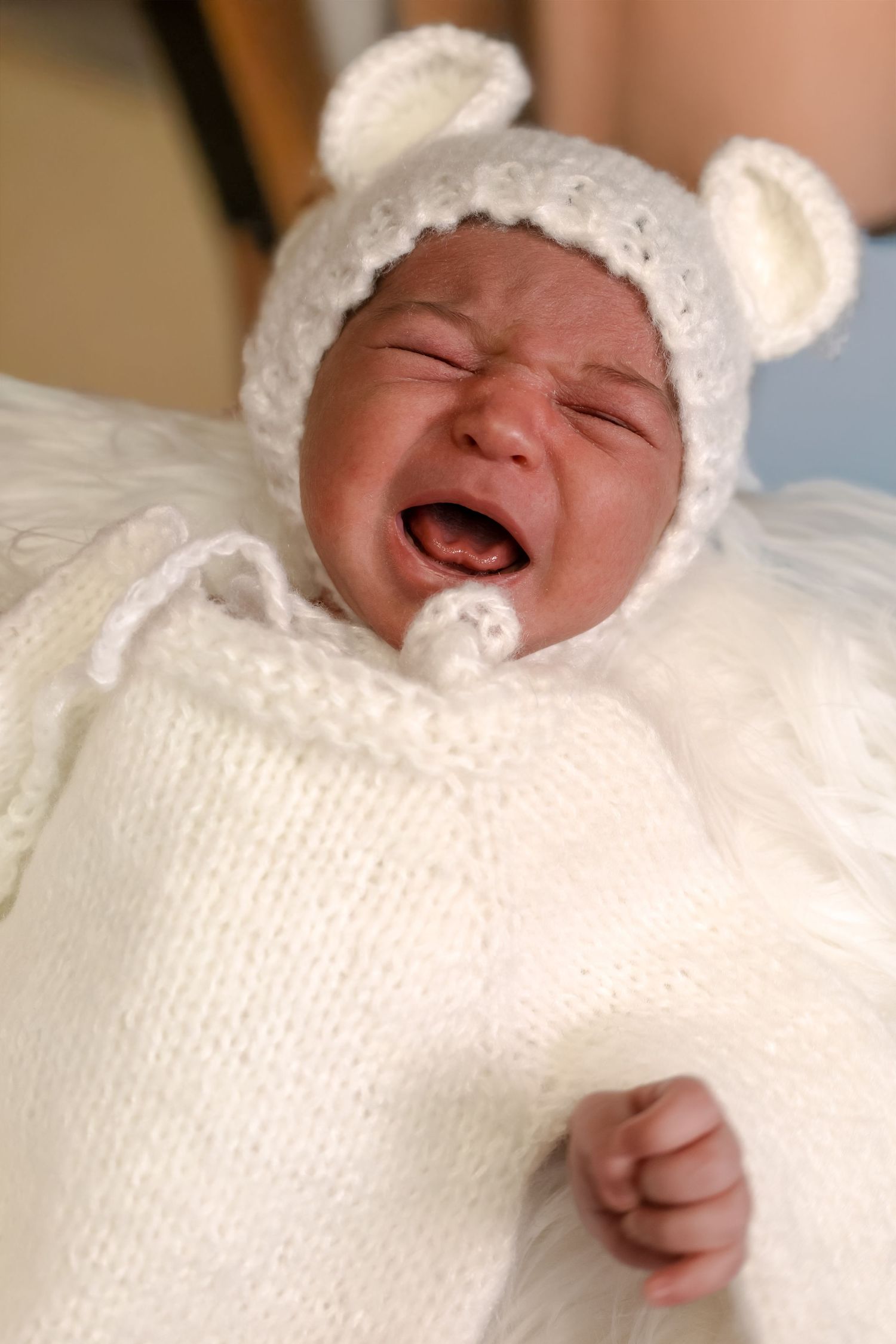 A crying baby wearing a white knit sweater and bear ear hat lies on white bedding.