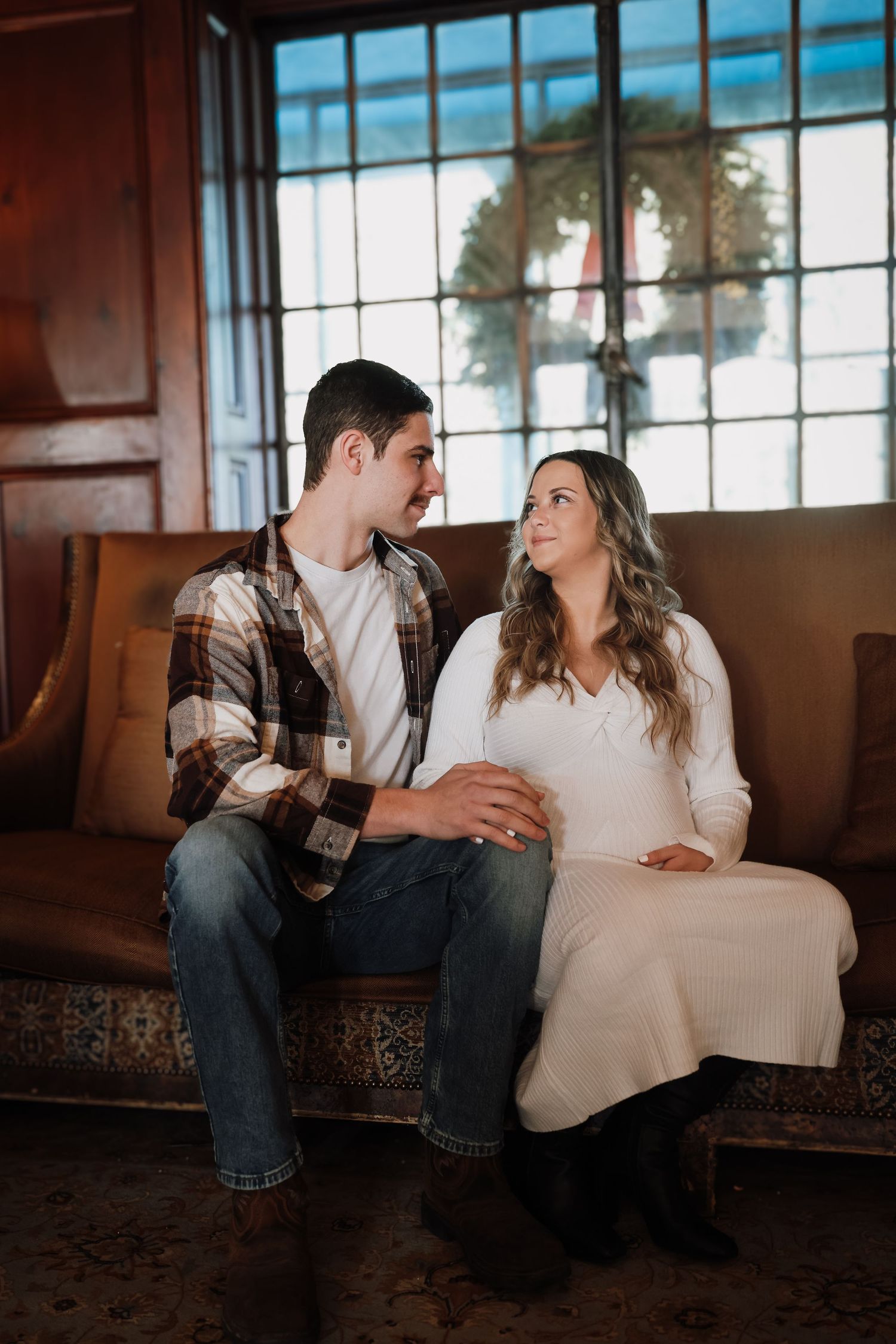 A couple shares an intimate moment while sitting together on a leather bench near large windows in a rustic setting.