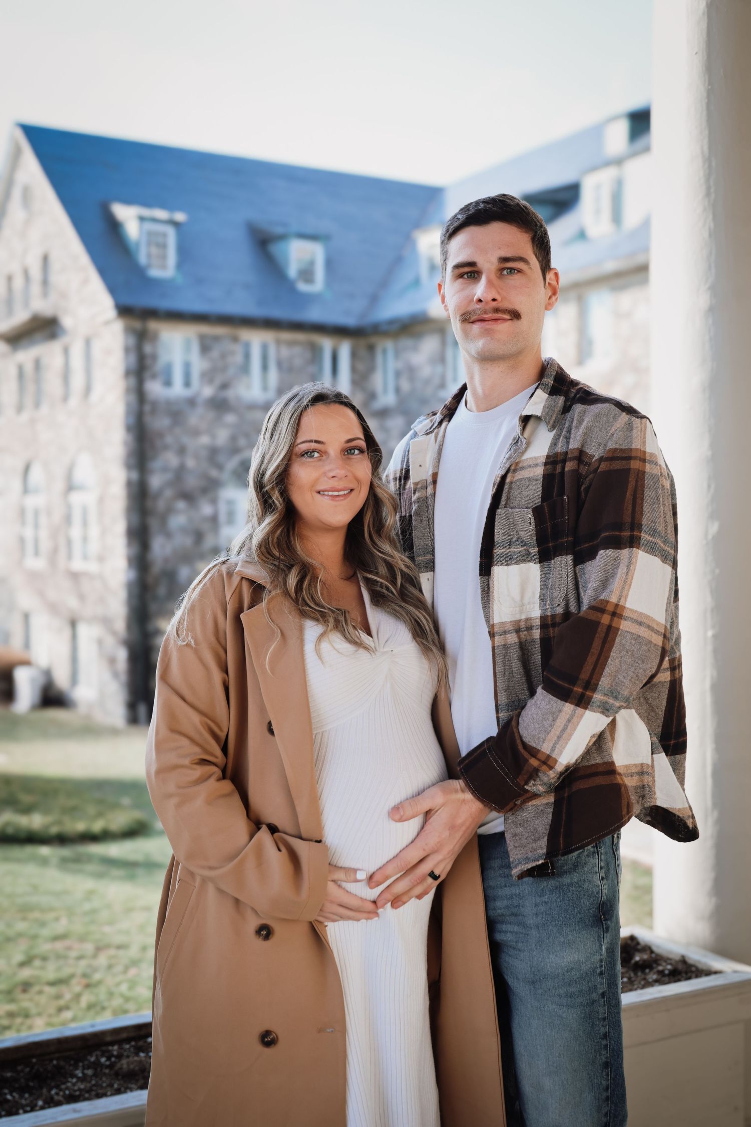 Couple poses together outdoors in front of stone building, wearing casual fall attire with camel coat and plaid shirt.