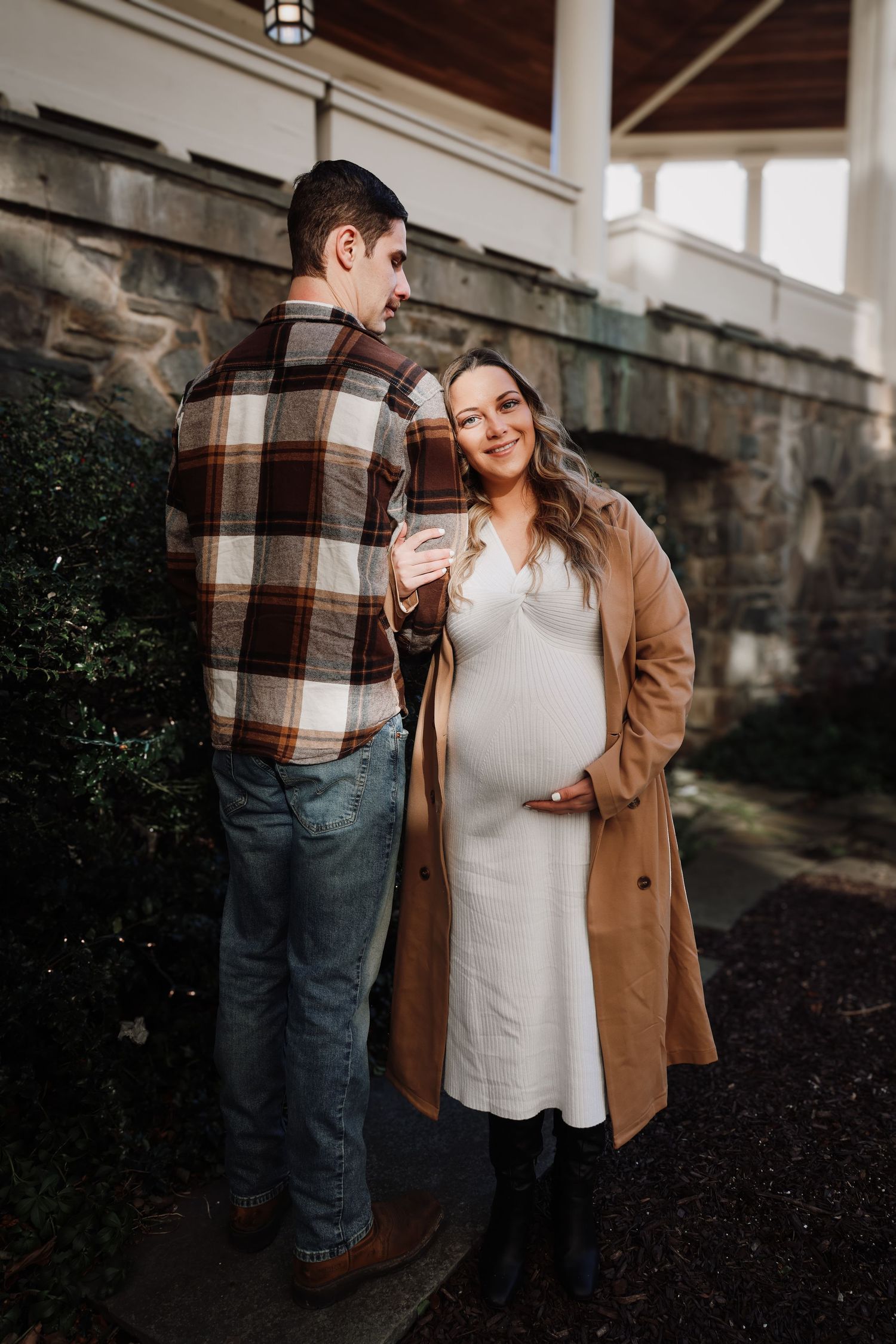 A couple shares a sweet moment outdoors, with the person in a tan coat showing a visible baby bump in a white dress.