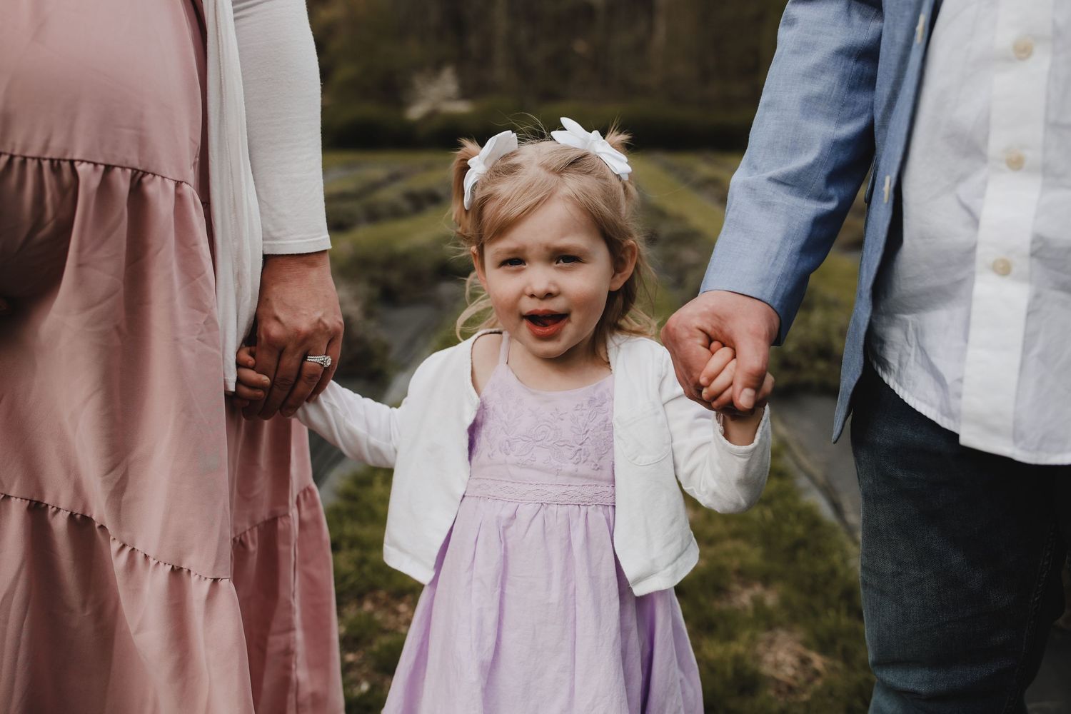 A small child in a lavender dress holds hands with adults while laughing joyfully outdoors.