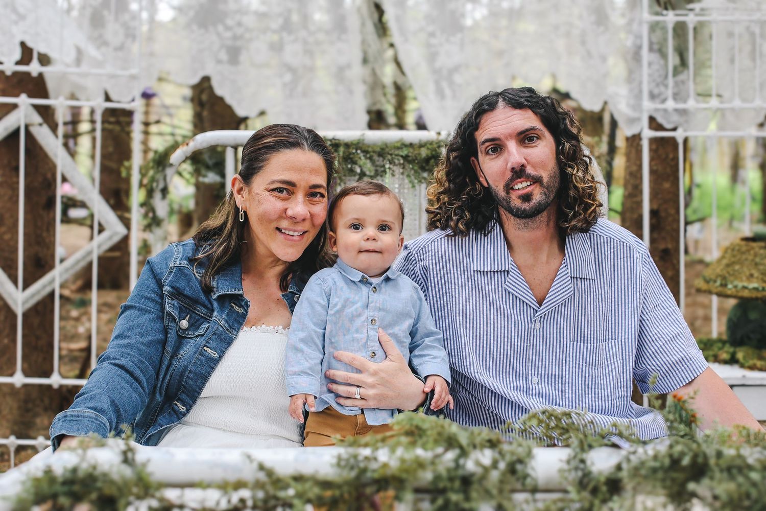 A family sits together outdoors on a rustic bench with greenery and wooden elements in the background.