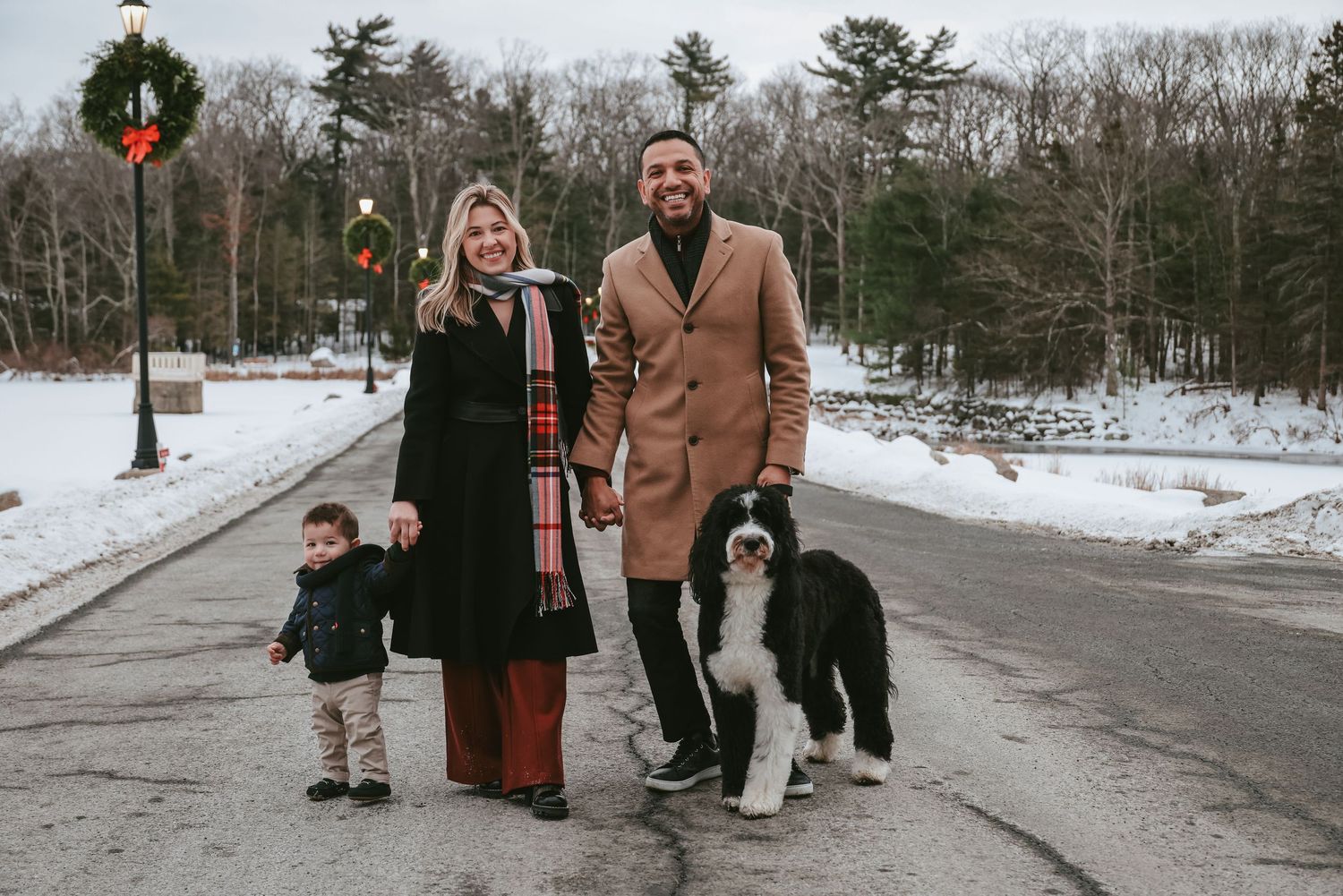 A family and their black and white dog stand together on a snowy winter road with evergreen trees in the background.