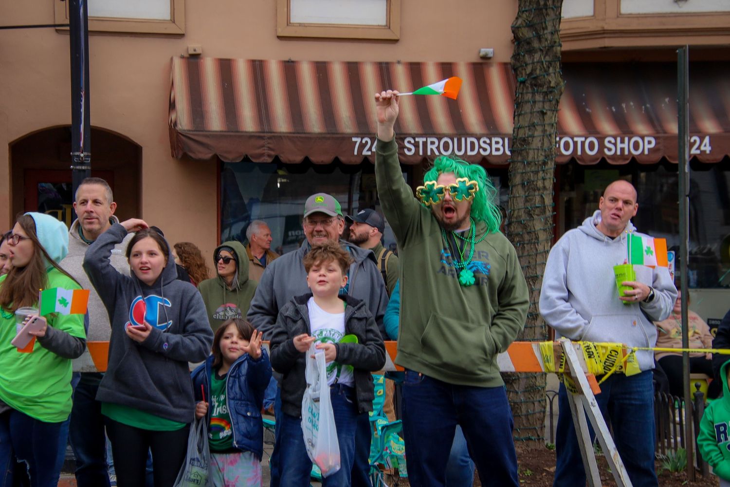 A crowd of spectators wearing green and white attire gathers along a street during a public event.