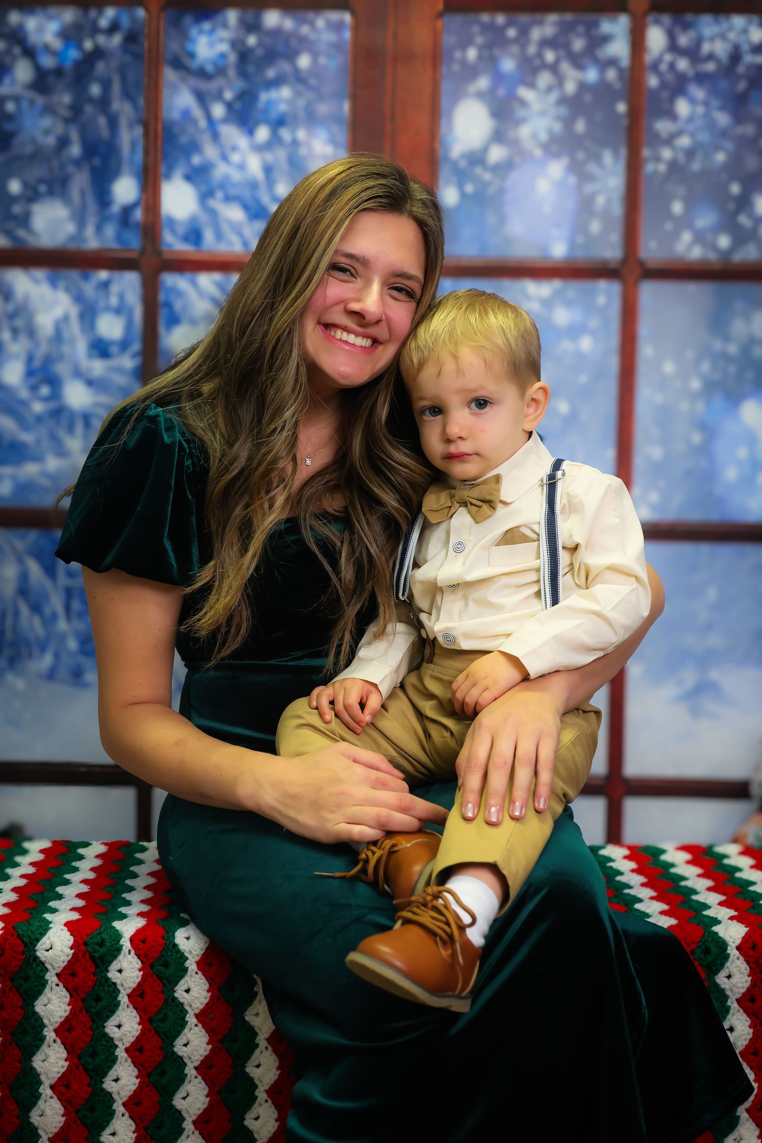 Mother and young child pose together on a festive holiday blanket with winter window backdrop.