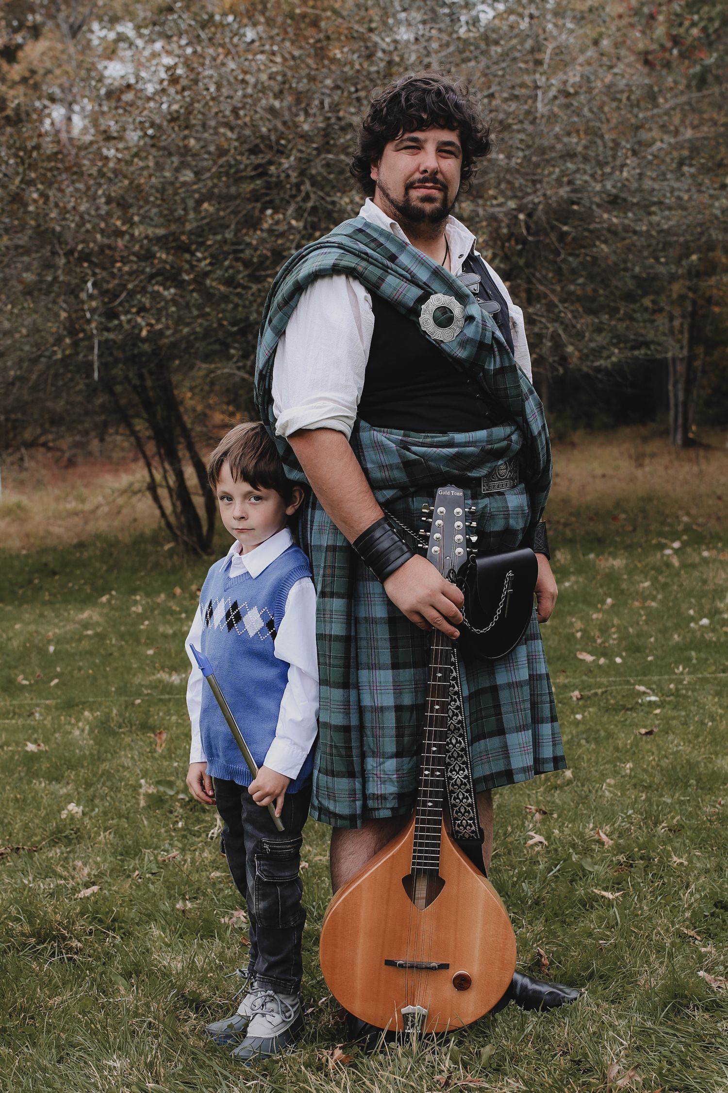 A parent and child in Scottish kilts stand together with a guitar in an autumn field.