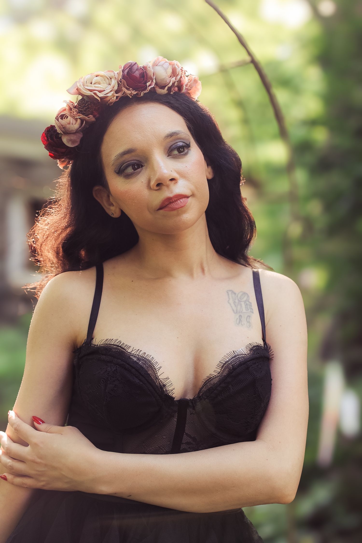 Woman in black lingerie poses outdoors with purple flowers in her hair against a natural backdrop.