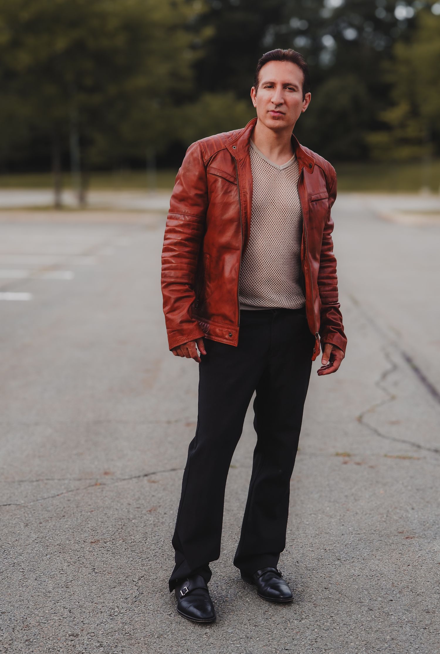 Person wearing brown leather jacket and dark pants stands on concrete surface in outdoor setting.