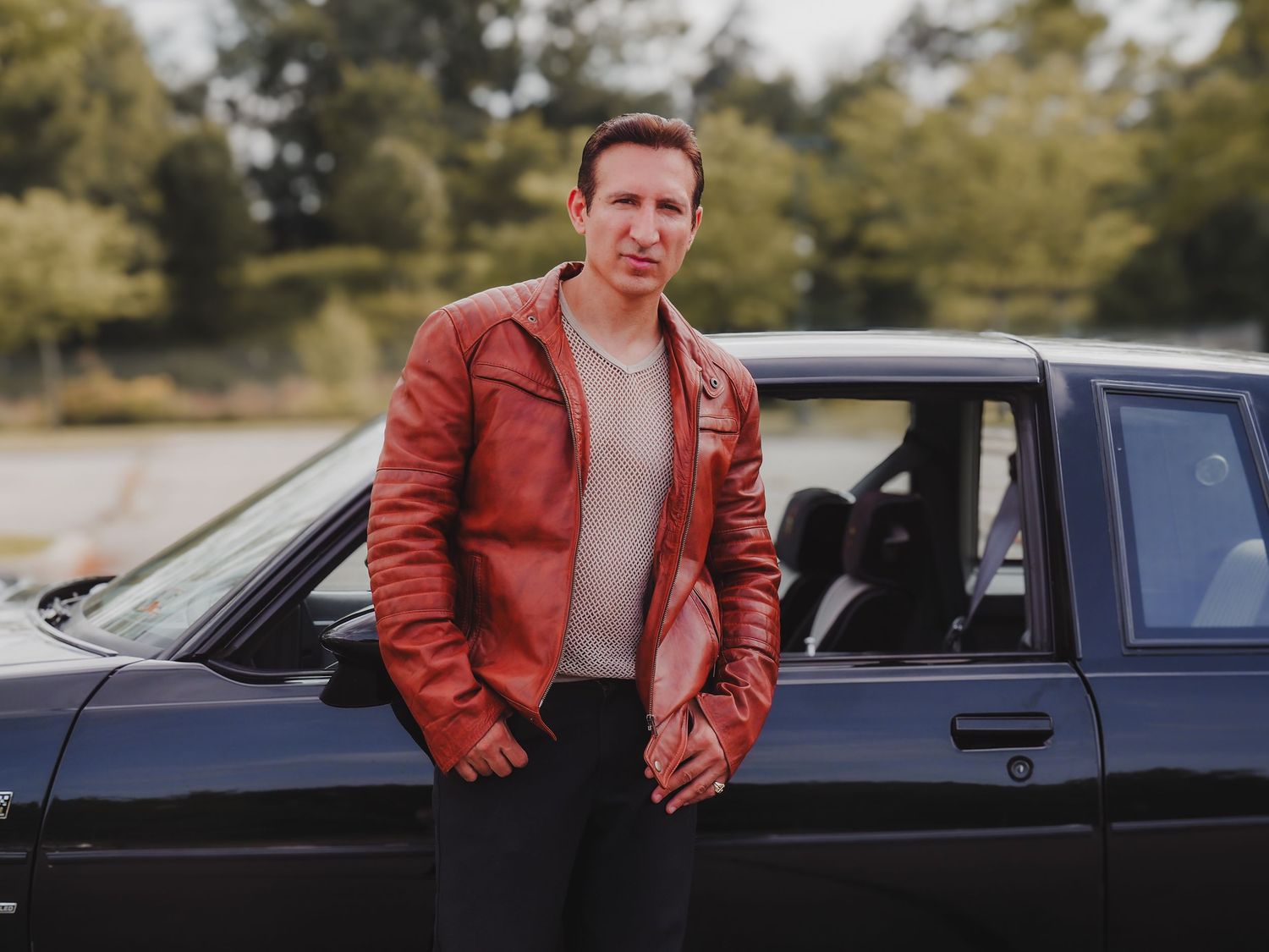A person in a red leather jacket stands next to a black classic car on a rural road at sunset.