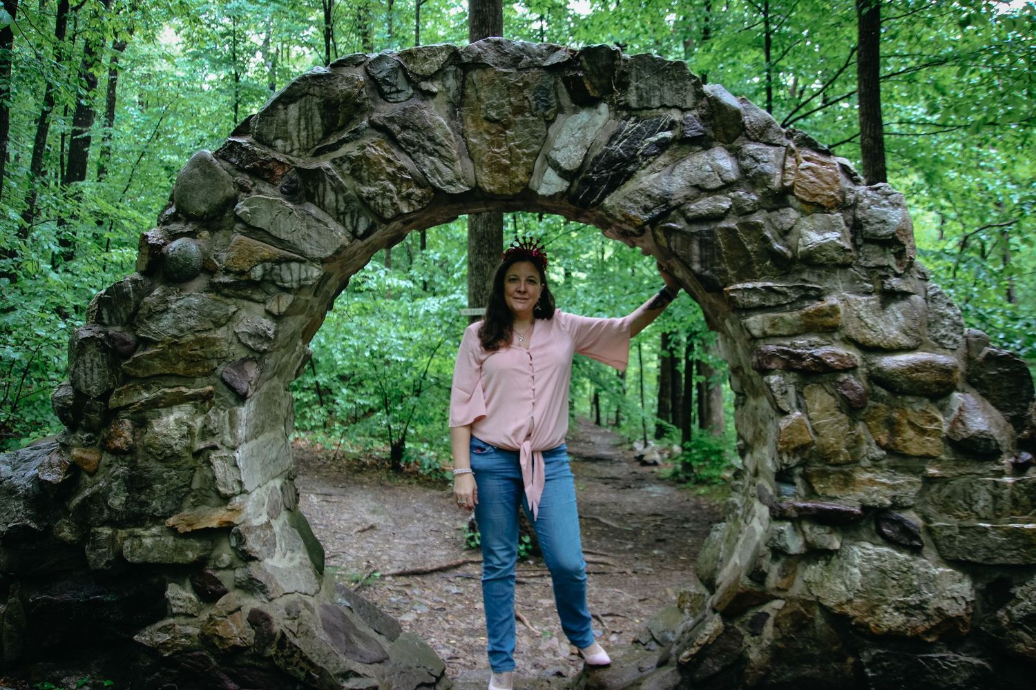A stone archway stands along a forest path surrounded by lush green trees and foliage.