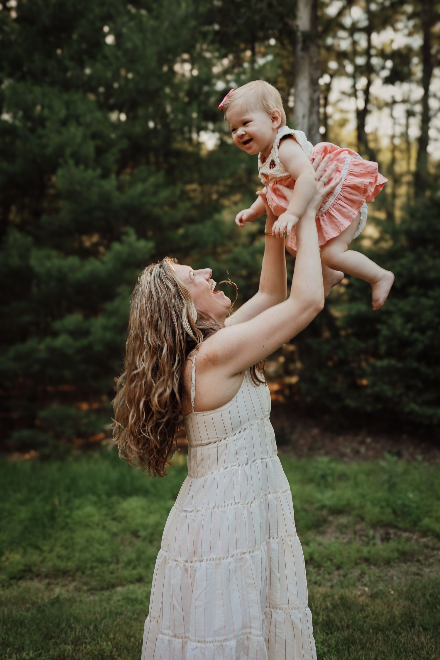 A person in a white dress lifts and plays with a baby outdoors in a natural wooded setting during golden hour.