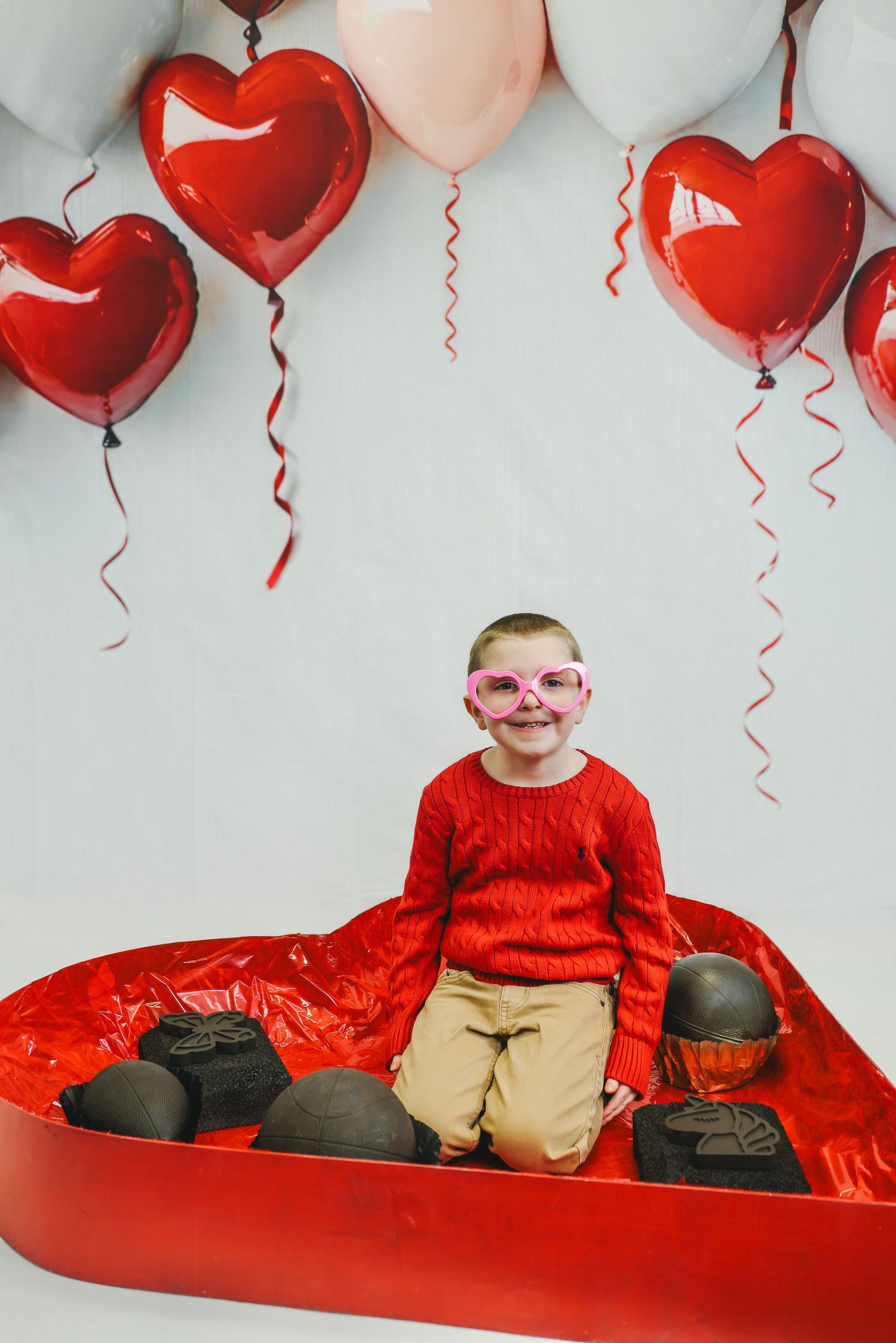 Valentine's day themed photo with red heart balloons and a red heart-shaped platform against white background.