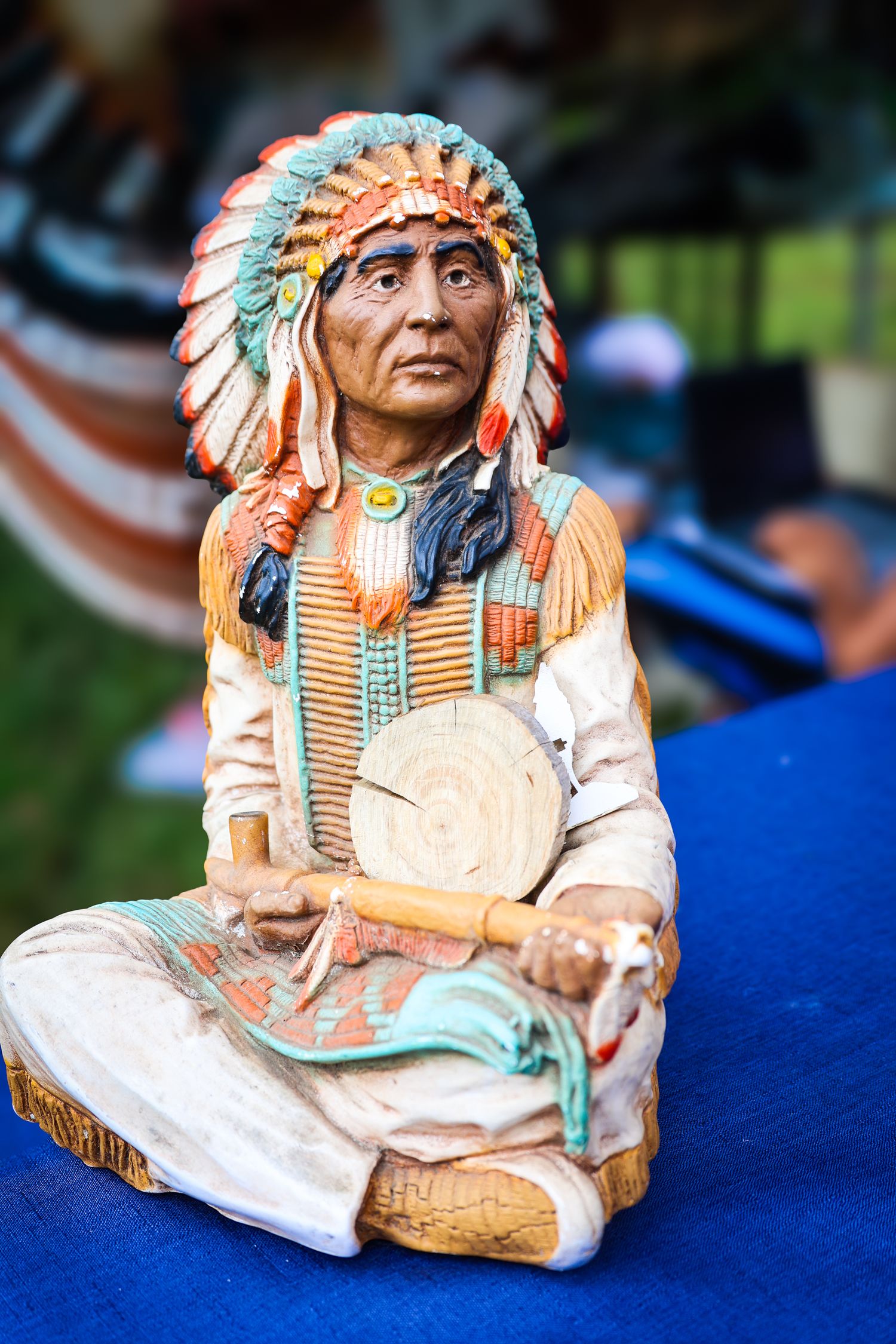 Ceramic figurine of a Native American chief wearing traditional headdress and white clothing displayed on blue fabric.