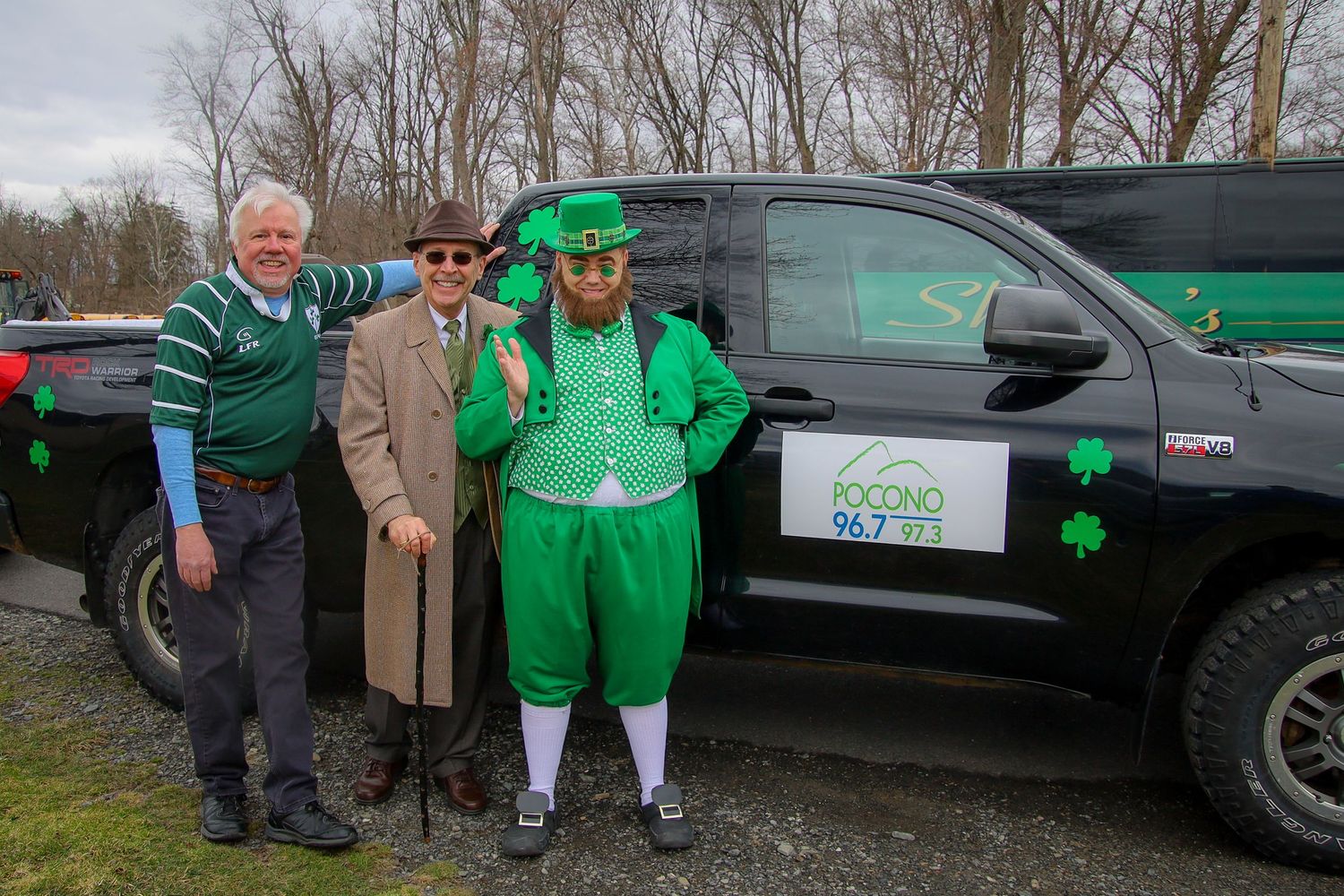A group of people wearing green clothing stand next to vehicles decorated with shamrocks for St. Patrick's Day.
